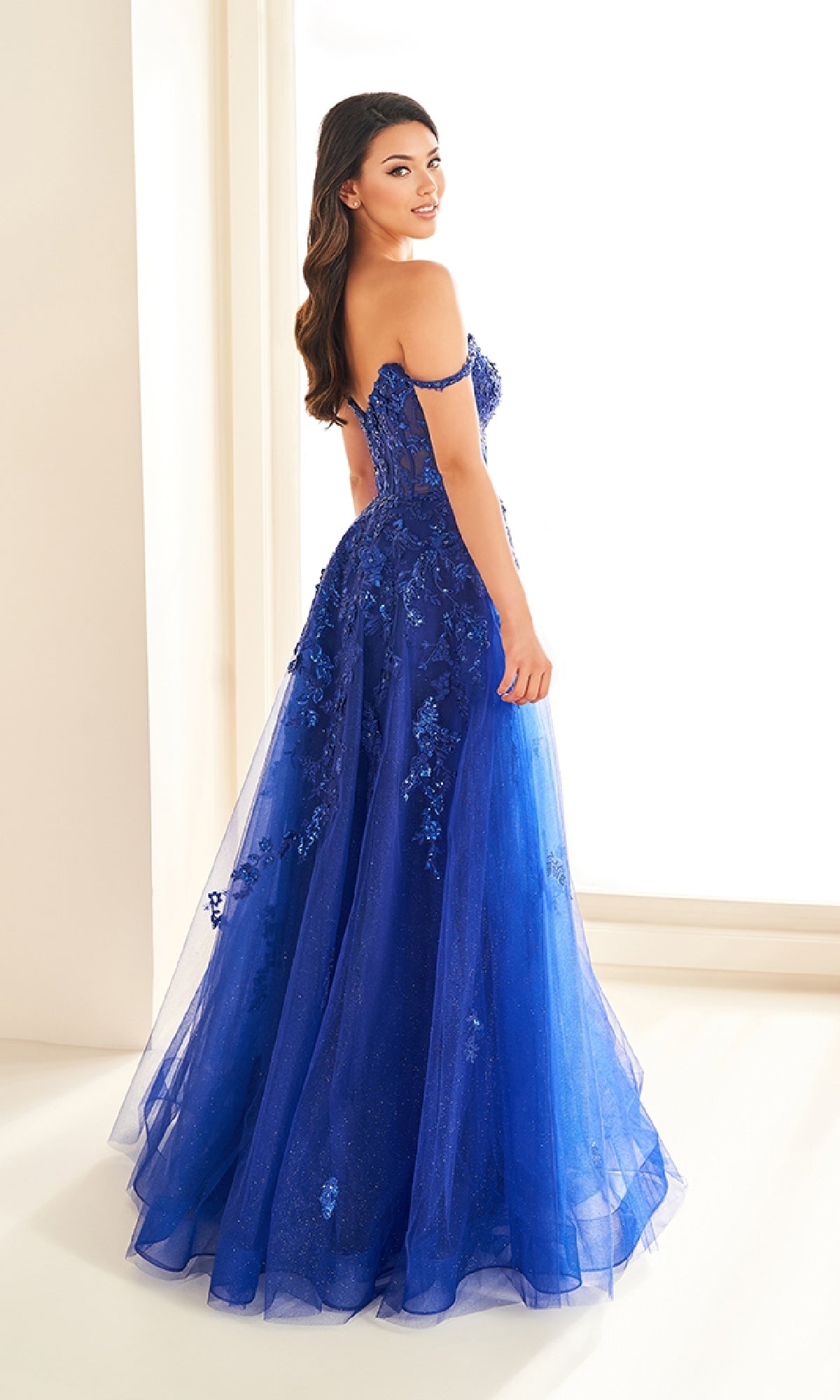 A woman stands indoors wearing the Ellie Wilde EW36204, a royal blue off-the-shoulder long prom dress with floral lace appliqués and a corset bodice, looking over her shoulder.