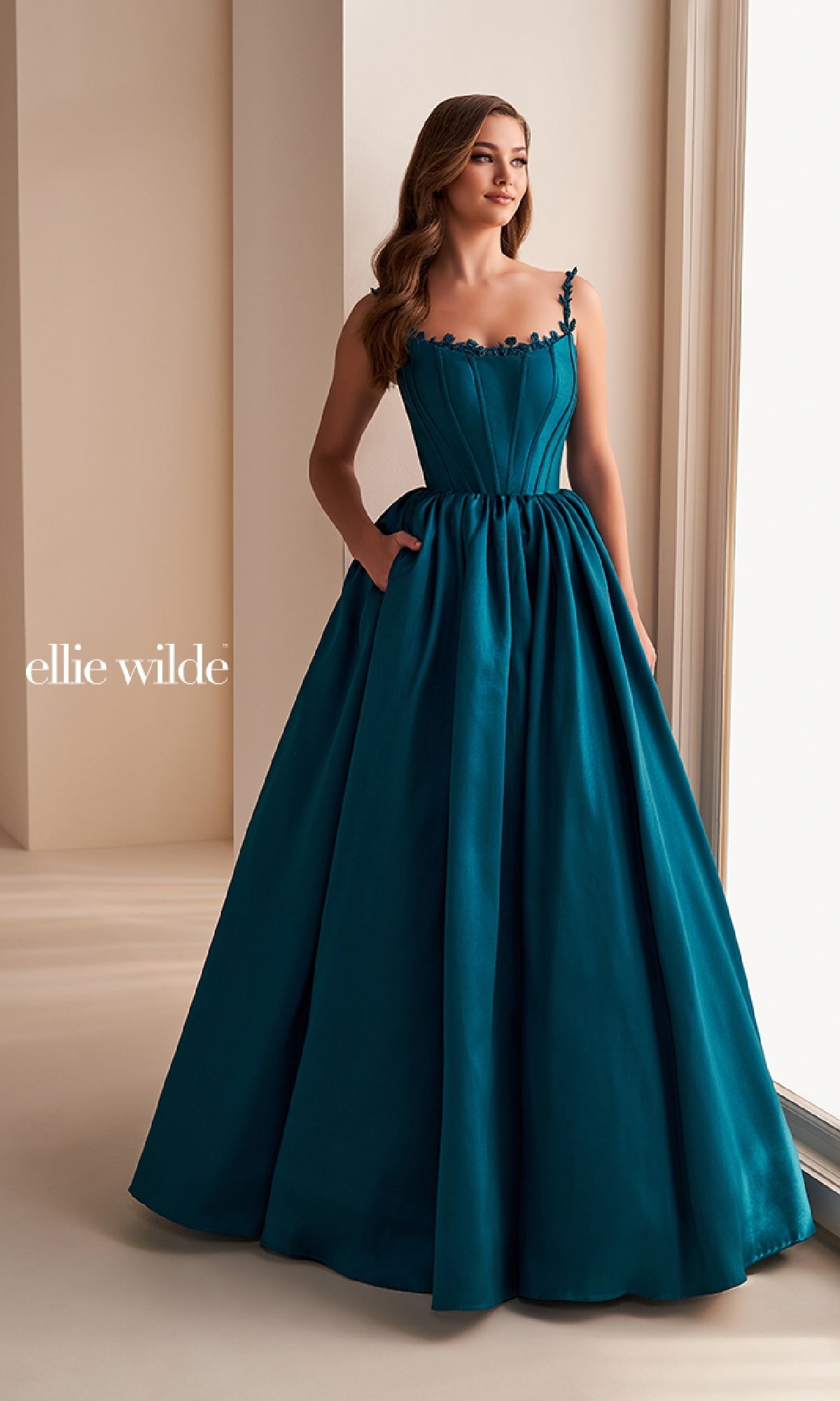 A woman stands near a window wearing the Ellie Wilde EW36221, a teal, sleeveless prom ball gown with a fitted corset bodice, full A-line skirt, and subtle floral details on the straps.