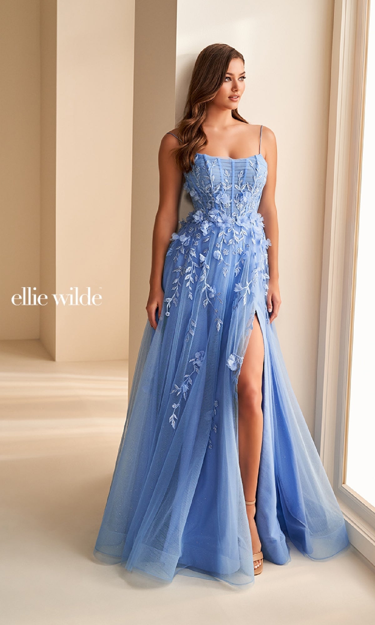 A woman stands by a window in a softly lit room wearing the Ellie Wilde EW36222, a light blue, floor-length prom dress with thin straps, lace applique, thigh-high slit, and fitted corset bodice.