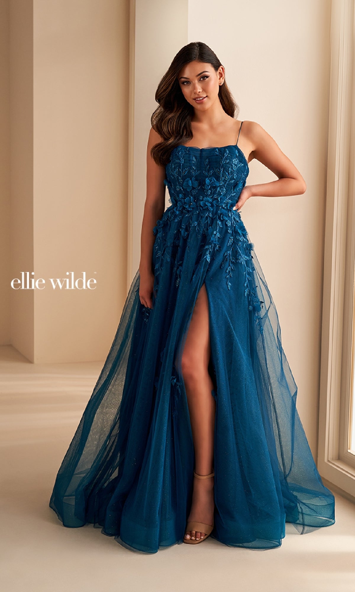 A woman wears the Ellie Wilde EW36222 long prom dress featuring teal lace applique, a sheer overlay, spaghetti straps, and a high front slit as she stands in a softly lit room.