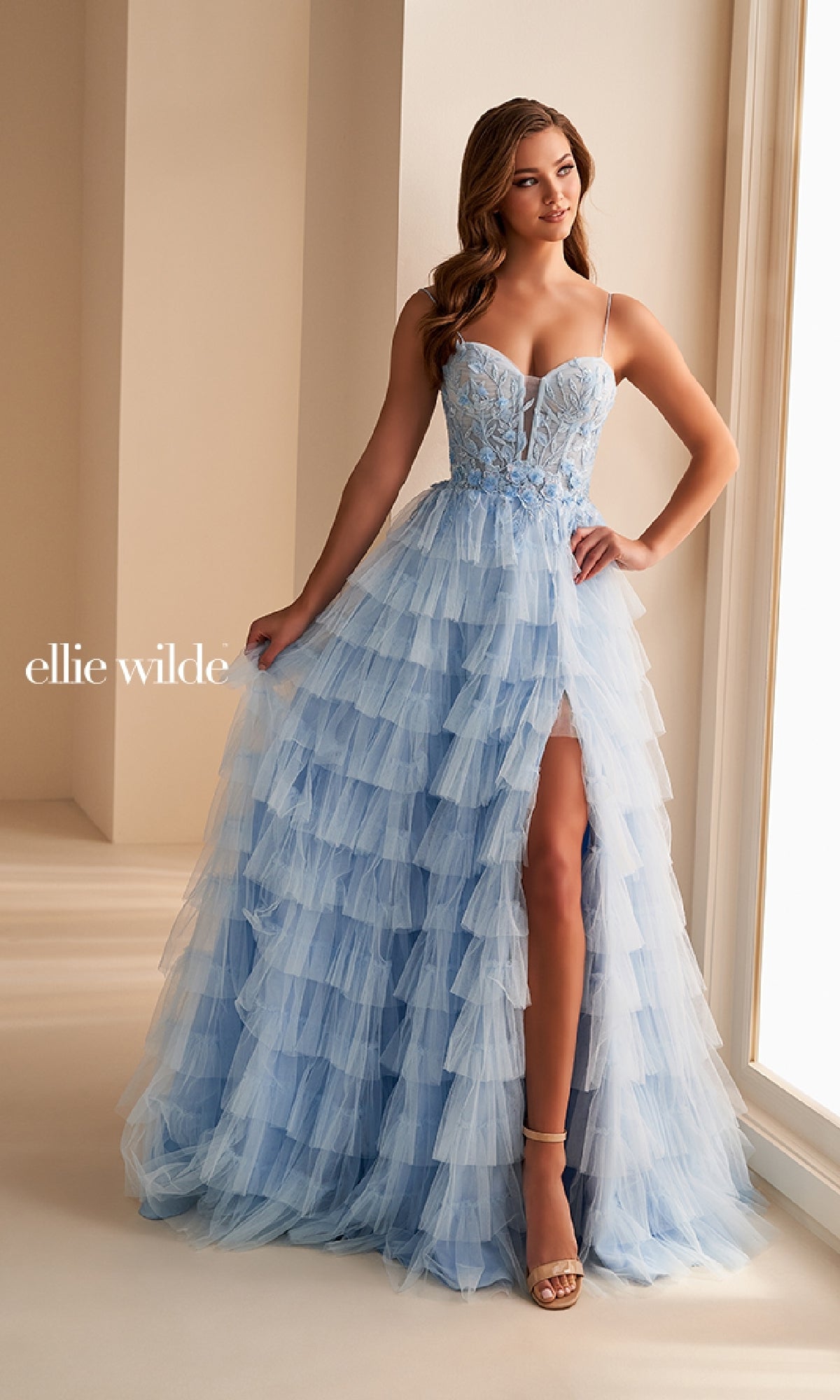 A woman stands indoors wearing the Ellie Wilde EW36223, a long prom dress featuring light blue tiered tulle, a fitted bodice, thin straps, and a thigh-high slit.