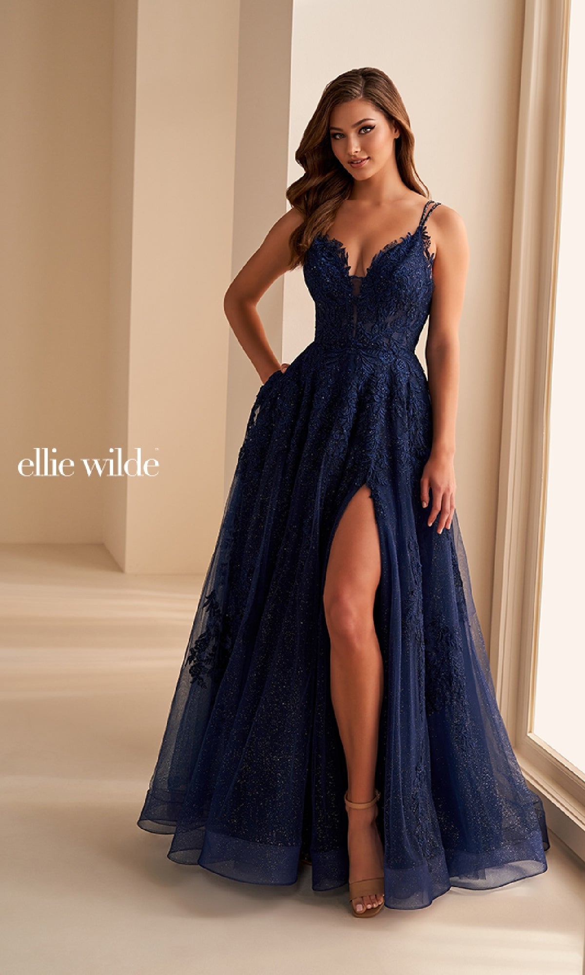 A woman stands indoors near a window wearing the Ellie Wilde EW36226, a deep blue floor-length A-line prom dress with a high slit and corset bodice.