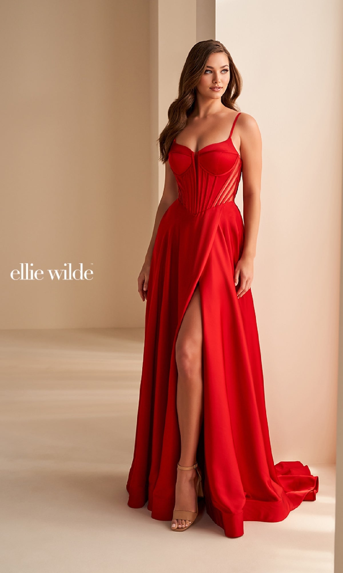 A woman stands indoors by a cream wall wearing the Ellie Wilde EW36227, a red, floor-length satin prom dress featuring an a-line silhouette, thigh-high slit, spaghetti straps, and open back.