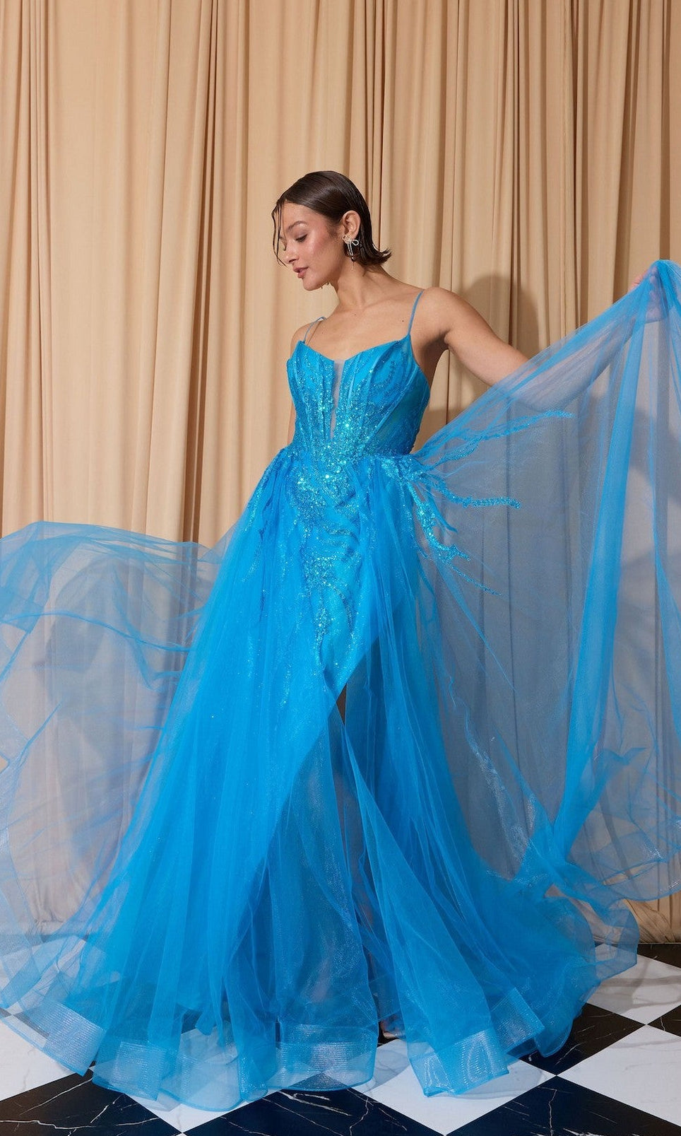 A woman in an azure blue Overskirt Long Sequin Prom Dress (Eureka EK112) with thin straps poses indoors by beige curtains, holding out the sparkling skirt of her gown.