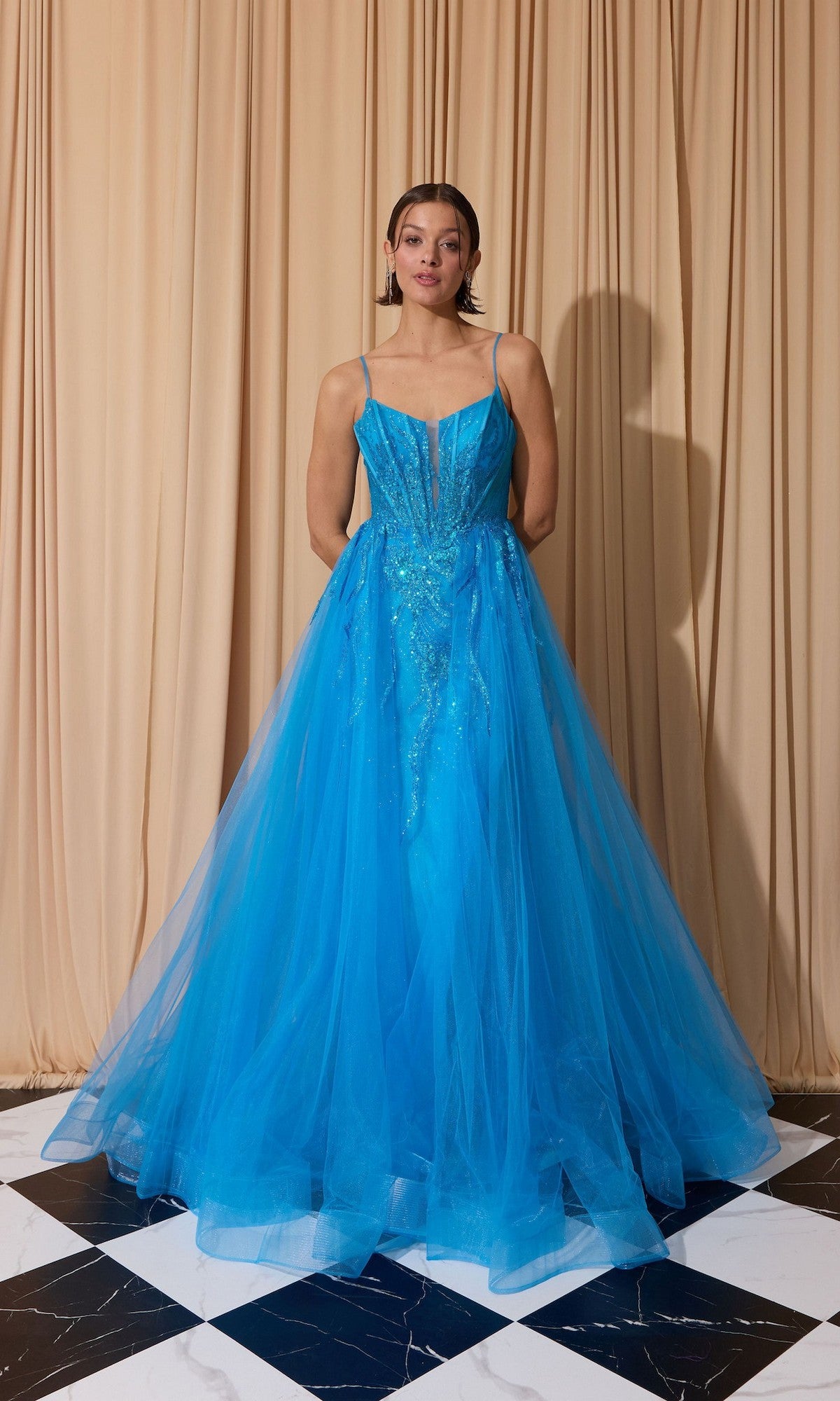 A woman stands on a black and white tiled floor in front of beige curtains, wearing the Eureka EK112 Overskirt Long Sequin Prom Dress featuring thin straps, a corset-style bodice, and sequin embellishments.