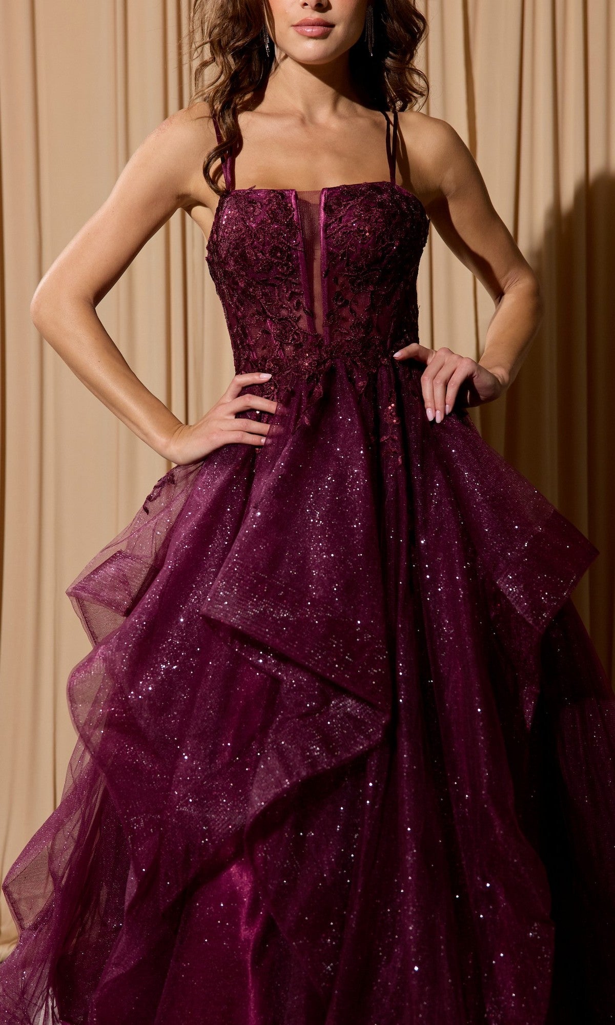 A woman stands against a beige curtain backdrop wearing the Eureka EK157 long prom dress—sleeveless, dark purple, featuring a glittery fitted bodice, lace-up open back, and layered tulle skirt.