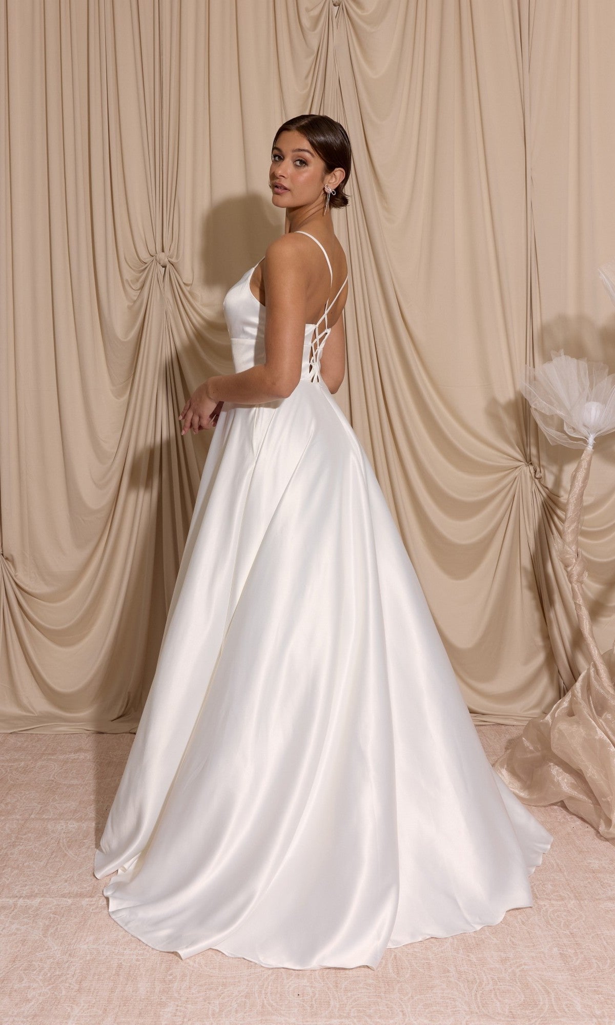 A woman in the Eureka EK190 Long Prom Dress, featuring a white satin a-line skirt and lace-up open back, stands before beige draped curtains, glancing over her shoulder.