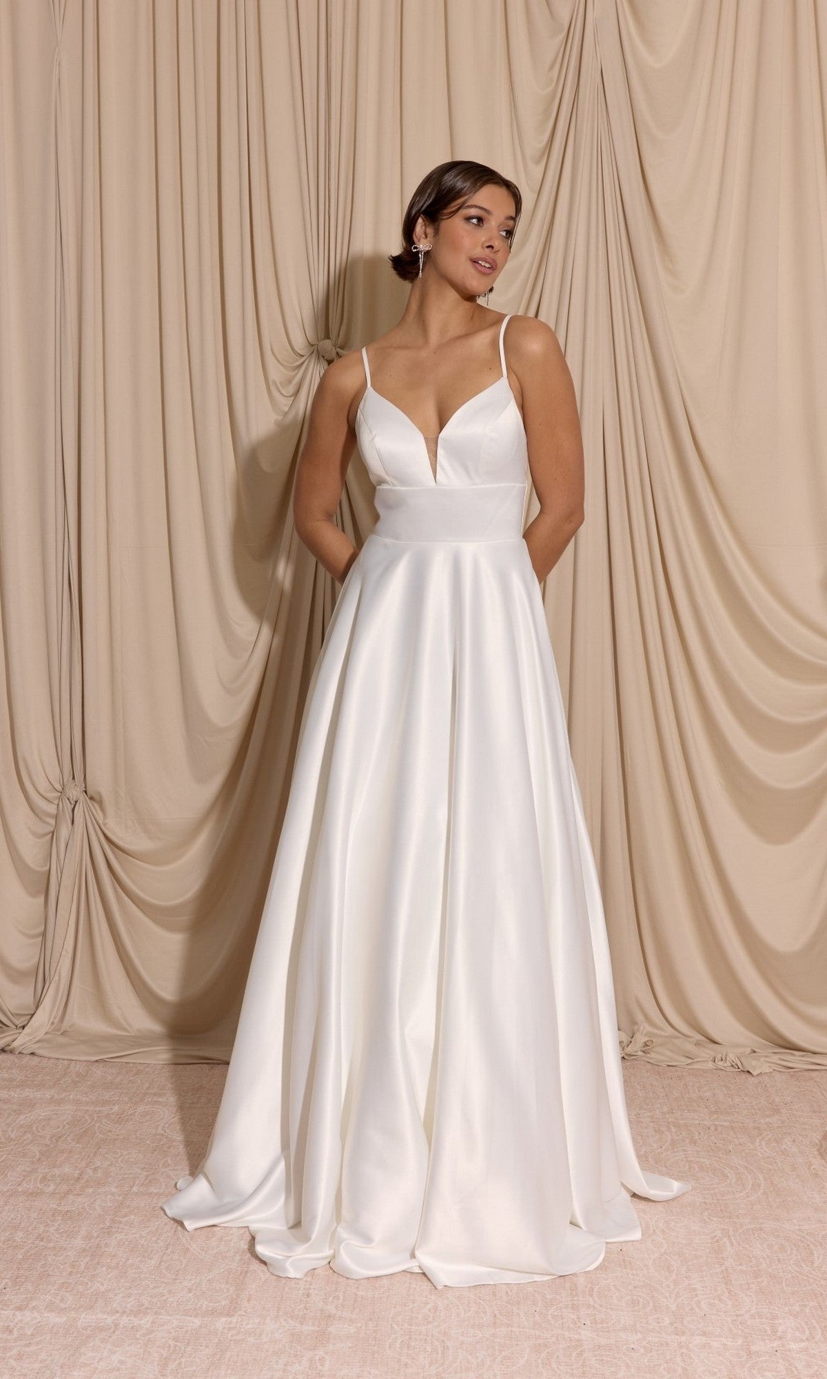 Woman wearing the Eureka EK190 long prom dress featuring a sleeveless white satin bodice, deep V-neckline, and a-line skirt, standing before beige draped fabric.