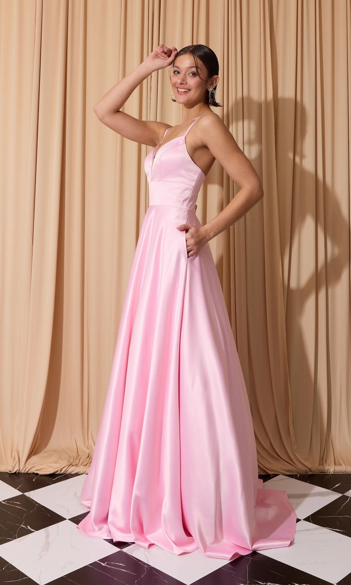 A woman poses on a black-and-white floor in front of beige curtains, wearing the Eureka EK190 long prom dress in light pink satin with thin straps and an open back.