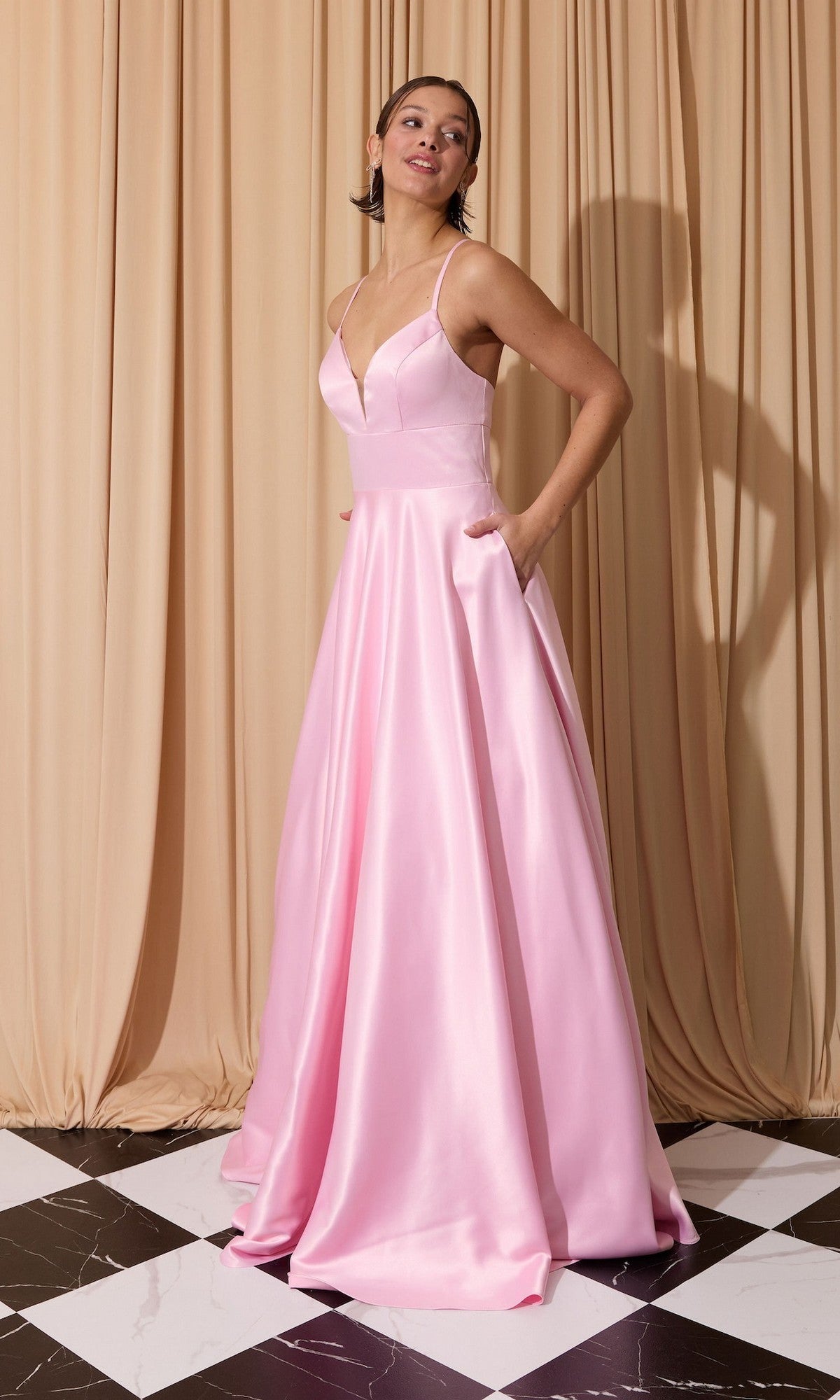 A woman stands on a black-and-white tiled floor, wearing the pink Eureka EK190 long prom dress with thin straps, open back, and pockets, posed in front of beige draped curtains.