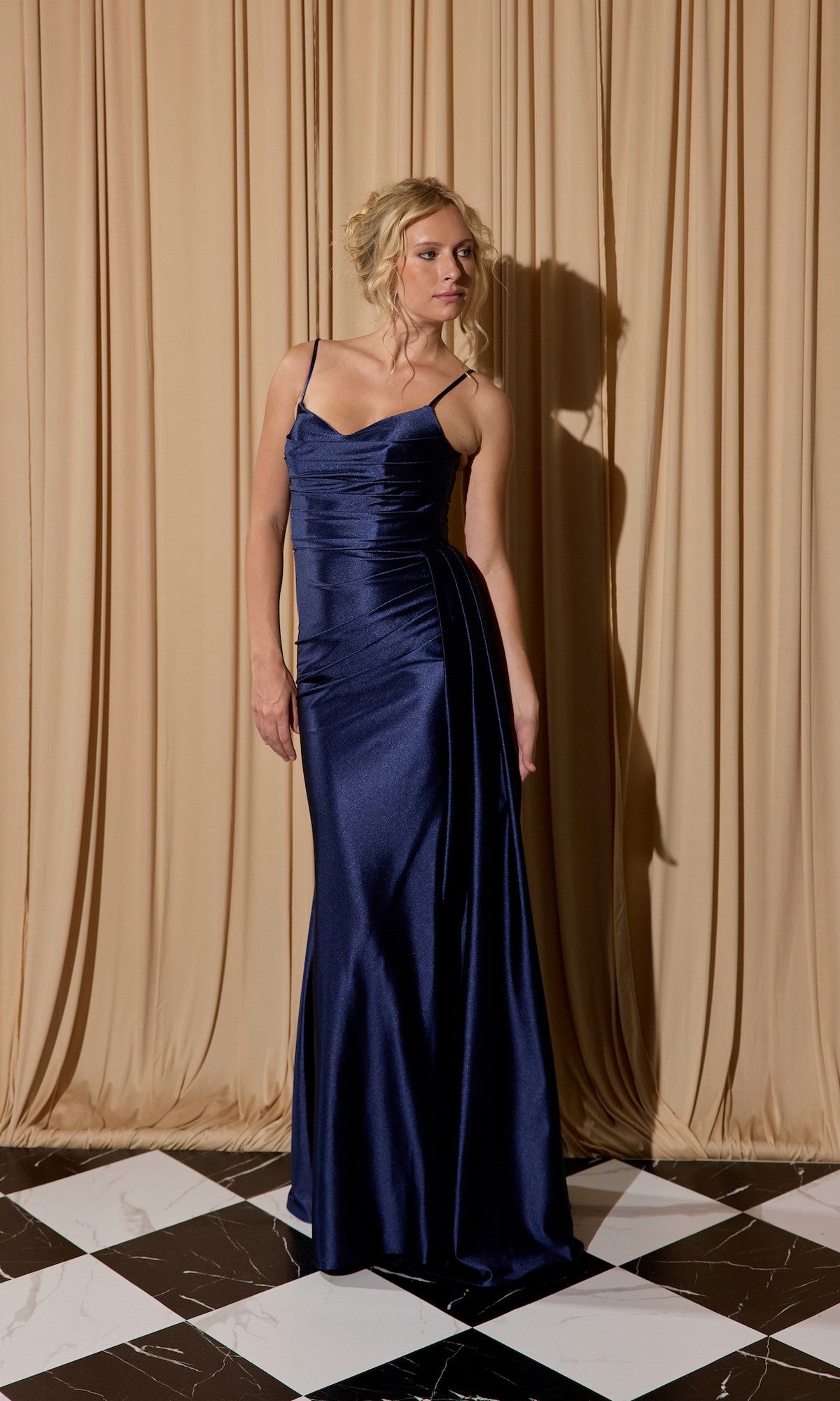 A woman in the Eureka EK199 Long Prom Dress in navy, featuring thin straps and a high-leg side slit, stands on a black and white tiled floor before beige curtains.