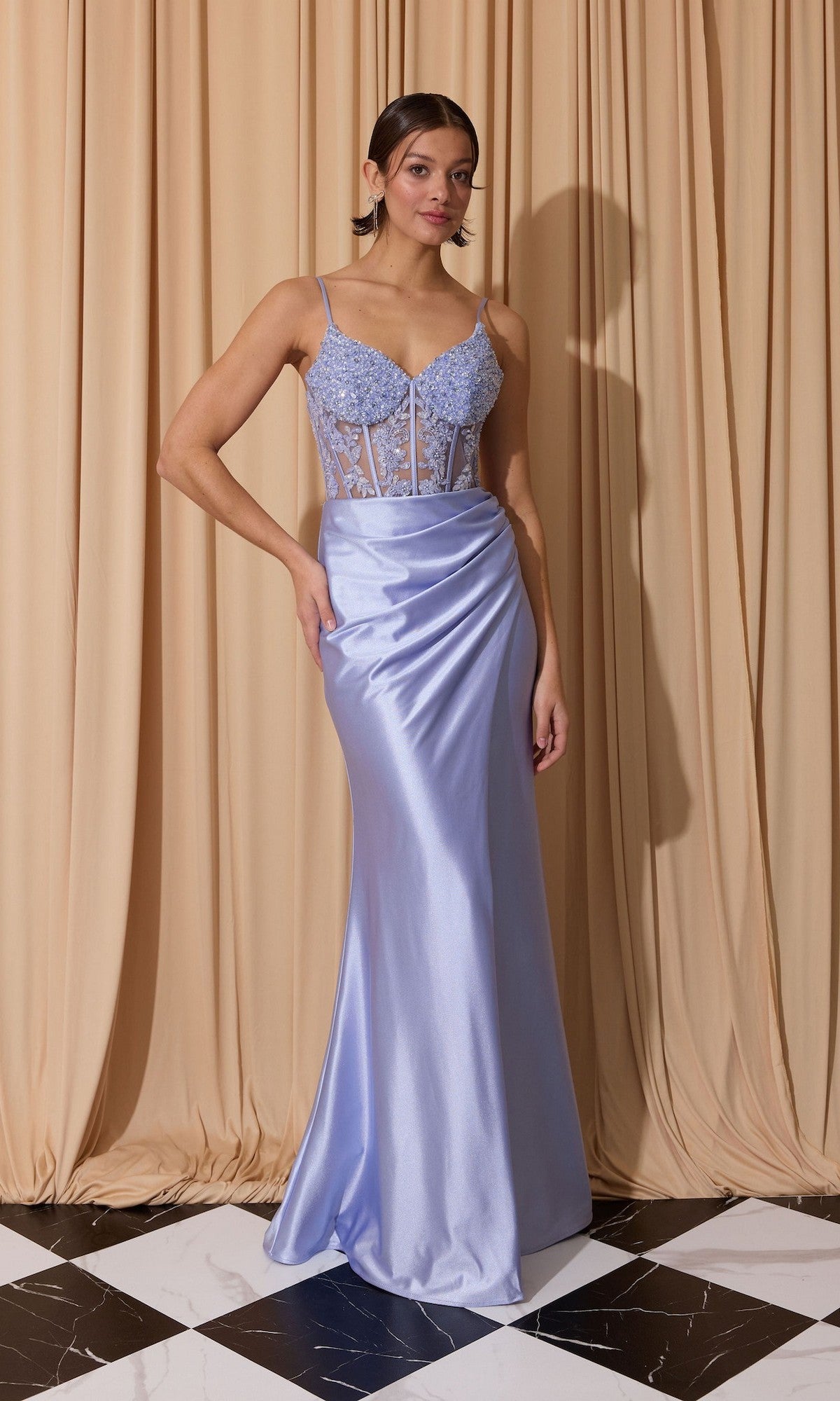 A woman stands on a black and white tiled floor in front of beige curtains, wearing the Eureka EK203 long prom dress—lavender, sleeveless, with a sheer corset bodice, lace details, and a graceful sheath skirt.