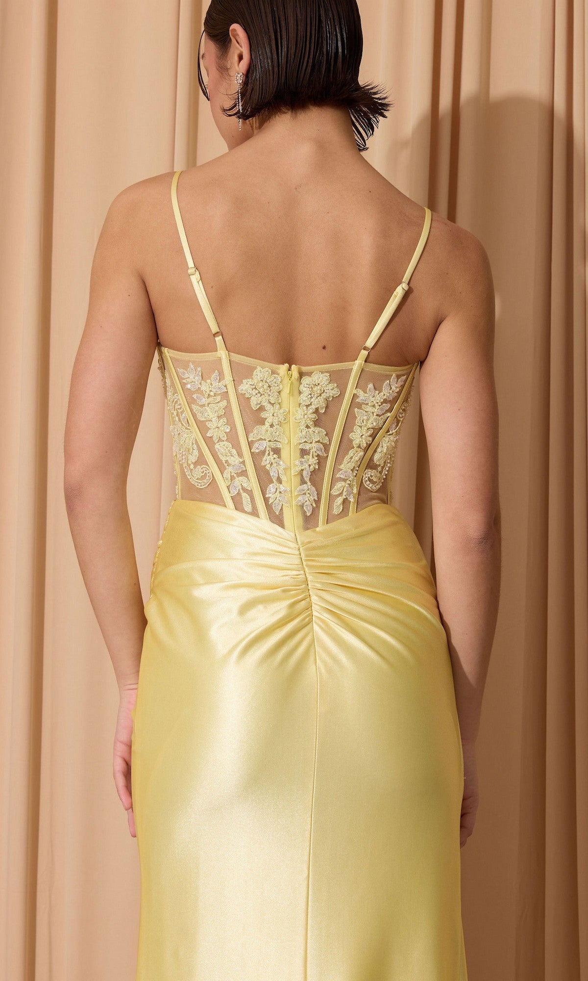 A person with short dark hair stands in front of beige curtains wearing the Eureka EK203 long prom dress—a yellow satin, sleeveless gown with a sheer embroidered back, corset bodice, and thin straps.