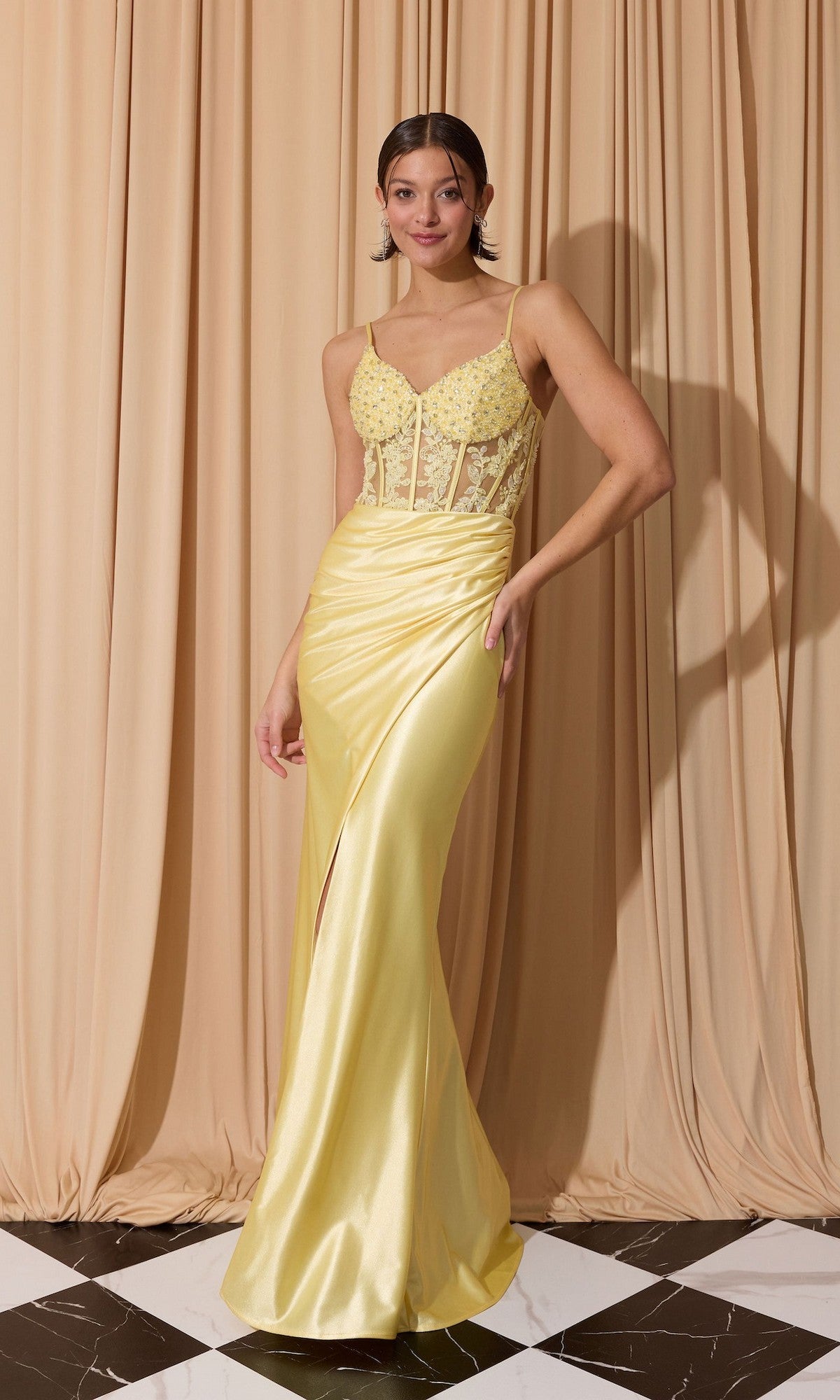 A woman poses in front of beige curtains on a black and white tiled floor, wearing the Eureka EK203 Long Prom Dress in Soft Yellow, featuring a satin corset bodice with spaghetti straps.
