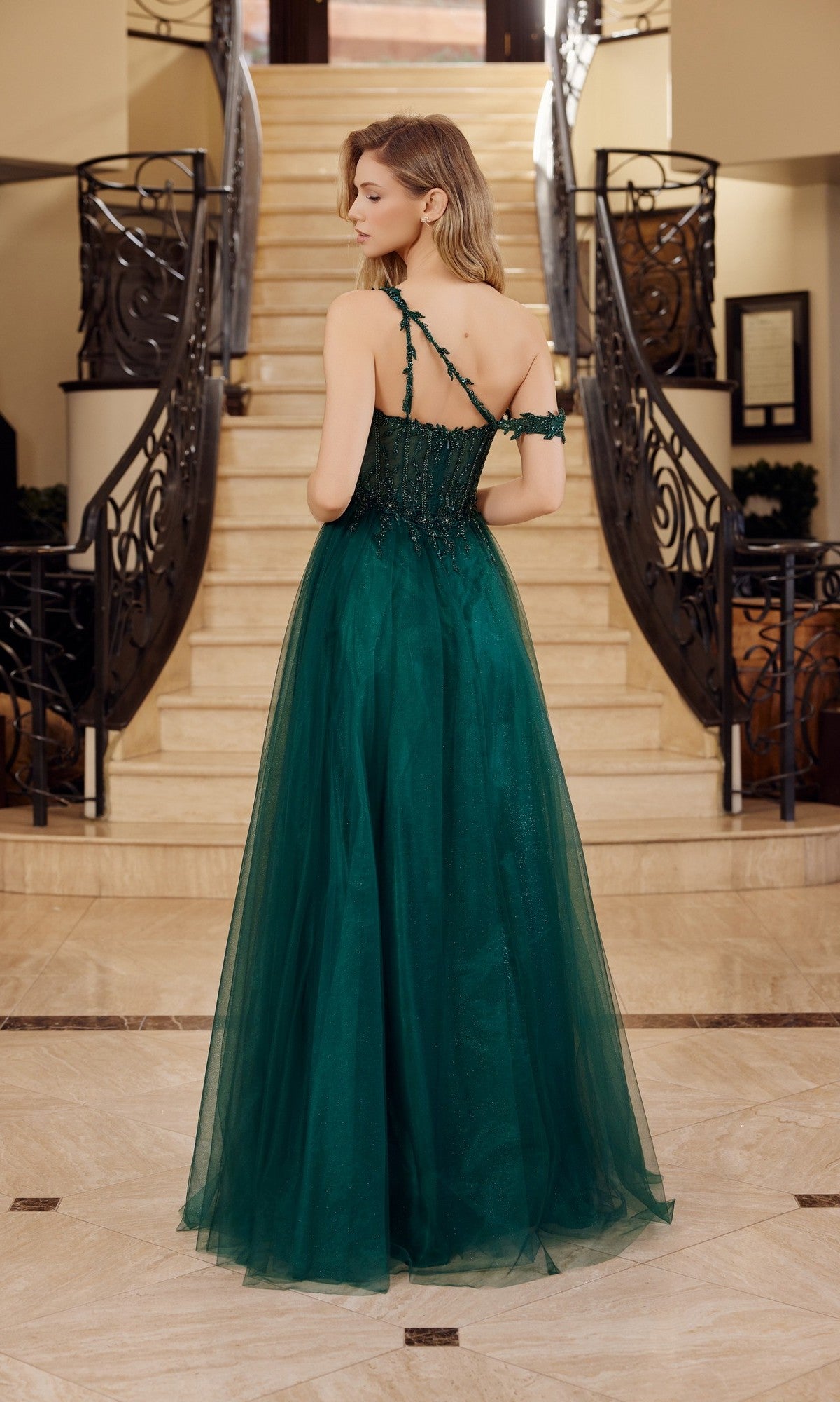 A woman in the Asymmetrical Glitter Prom Dress: Nox Anabel F1567 stands with her back to the camera at the bottom of a staircase with ornate railings.