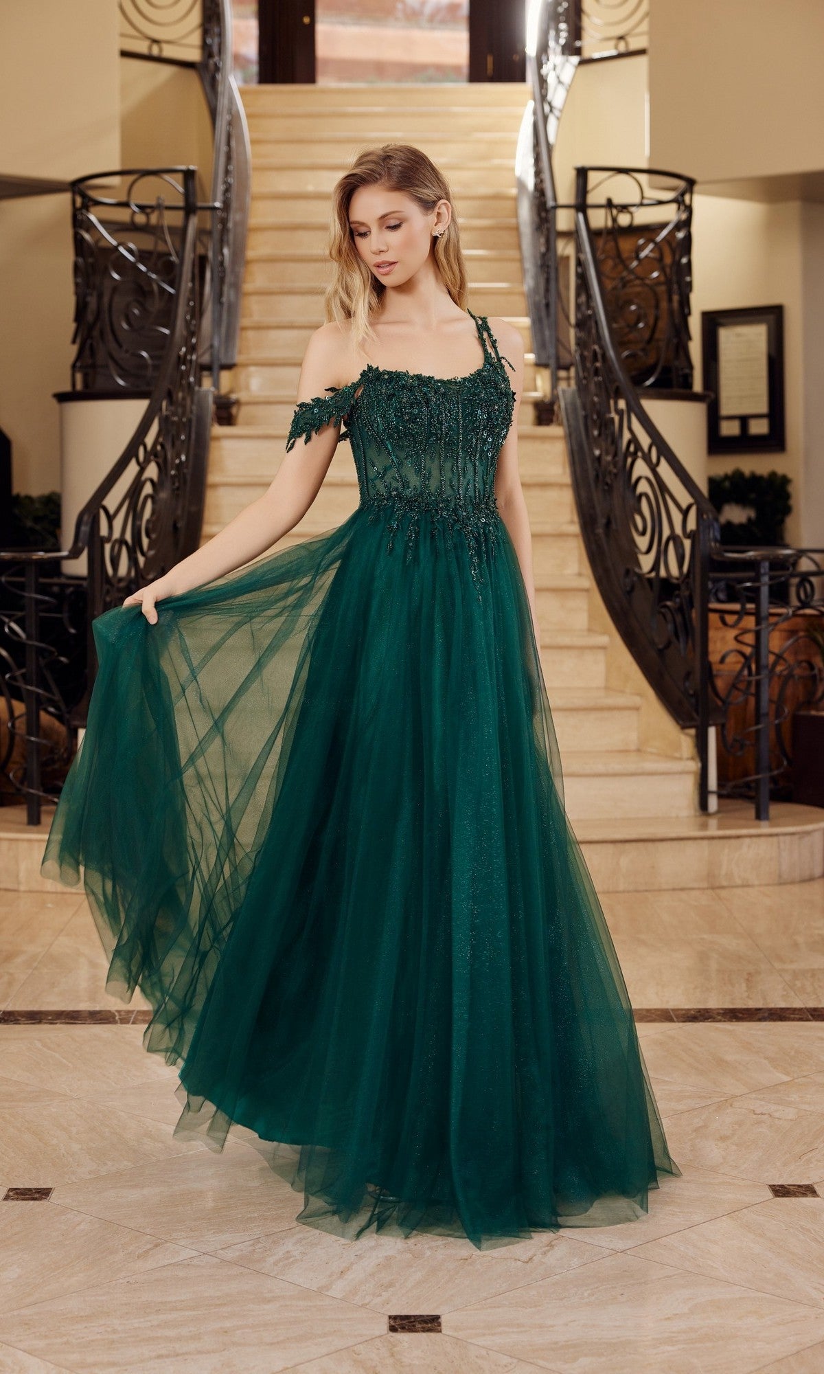 A woman stands by a staircase, holding out the side of her off-the-shoulder, floor-length Nox Anabel F1567 Asymmetrical Glitter Prom Dress in emerald.