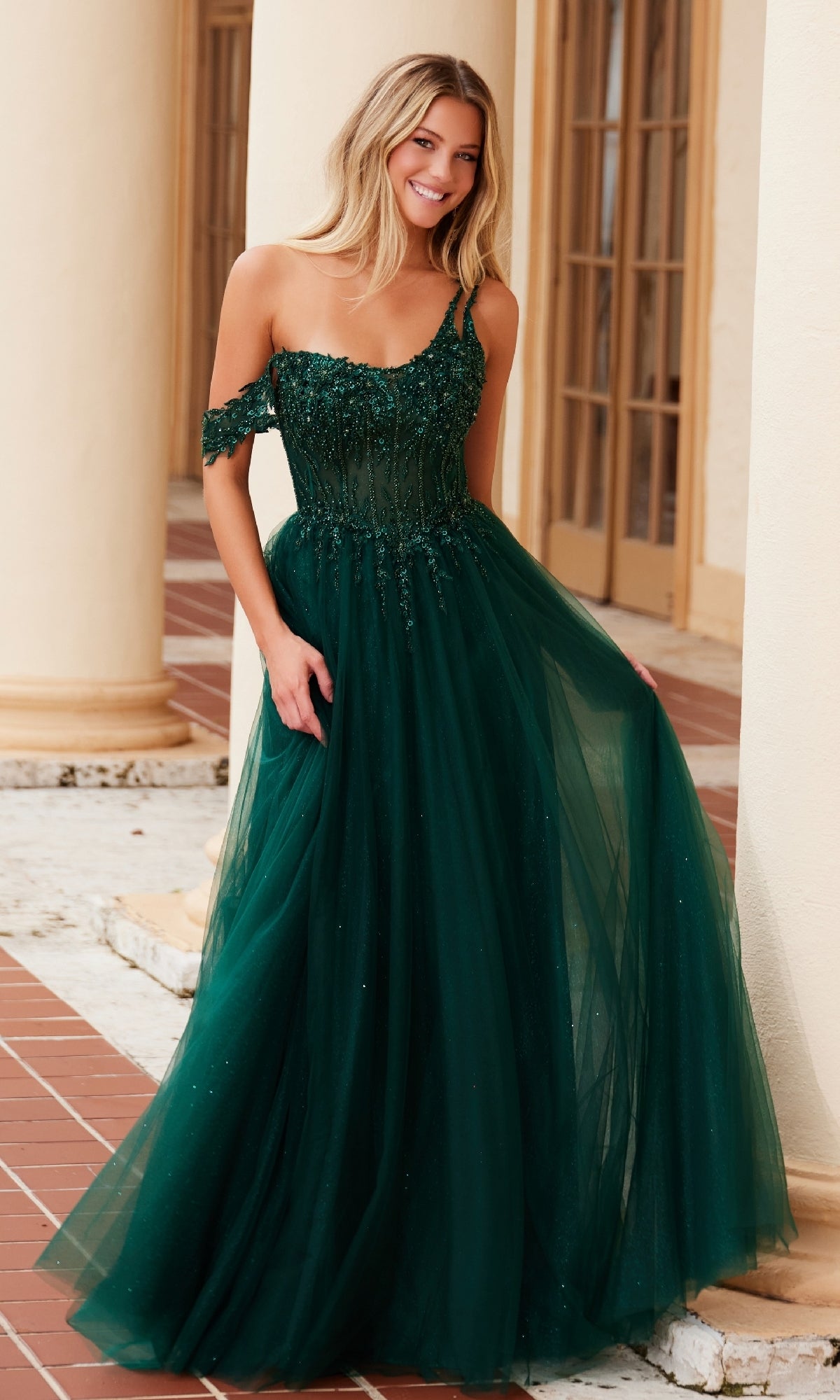 A woman stands outdoors wearing the Nox Anabel F1567, an asymmetrical glitter prom dress with a fitted bodice and full tulle skirt.
