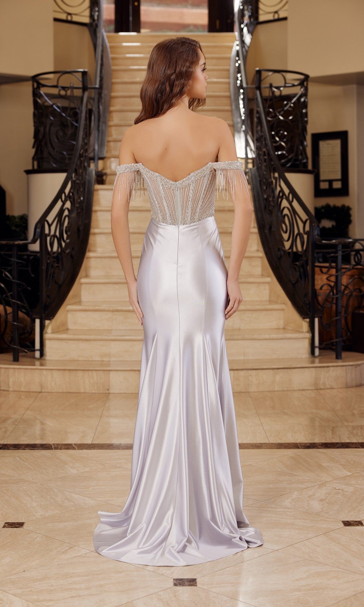 A woman in the Fringed-Bodice Long Prom Dress: Nox Anabel F1568, a strapless, floor-length silver satin gown with intricate beading, stands at the base of a staircase, facing away from the camera.