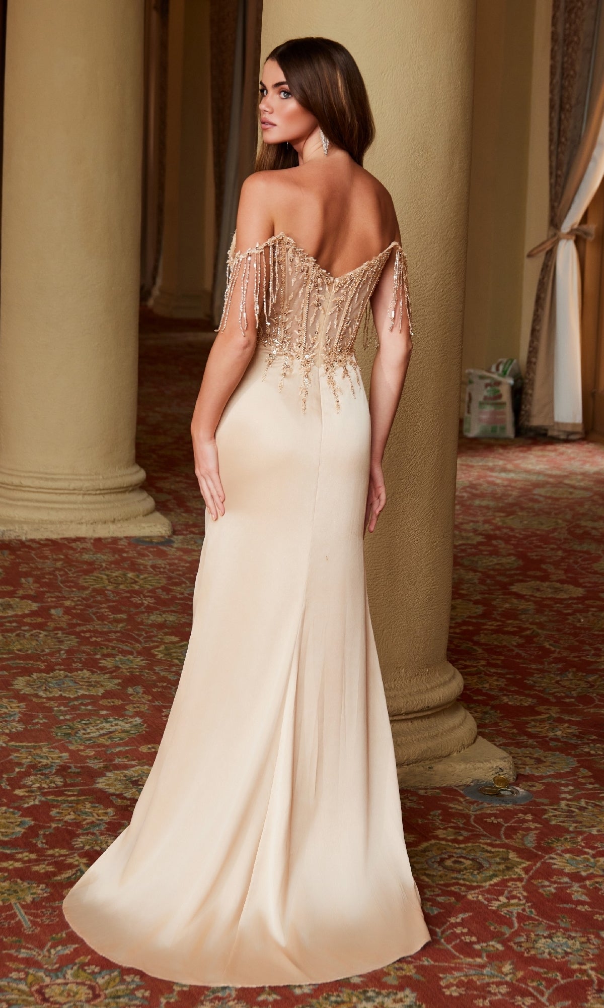 A woman stands with her back to the camera in the Nox Anabel F1571 Beaded-Bodice Long Prom Dress, showcasing off-the-shoulder style, a beaded bodice, and fringe details in an ornate room with columns and patterned carpet.