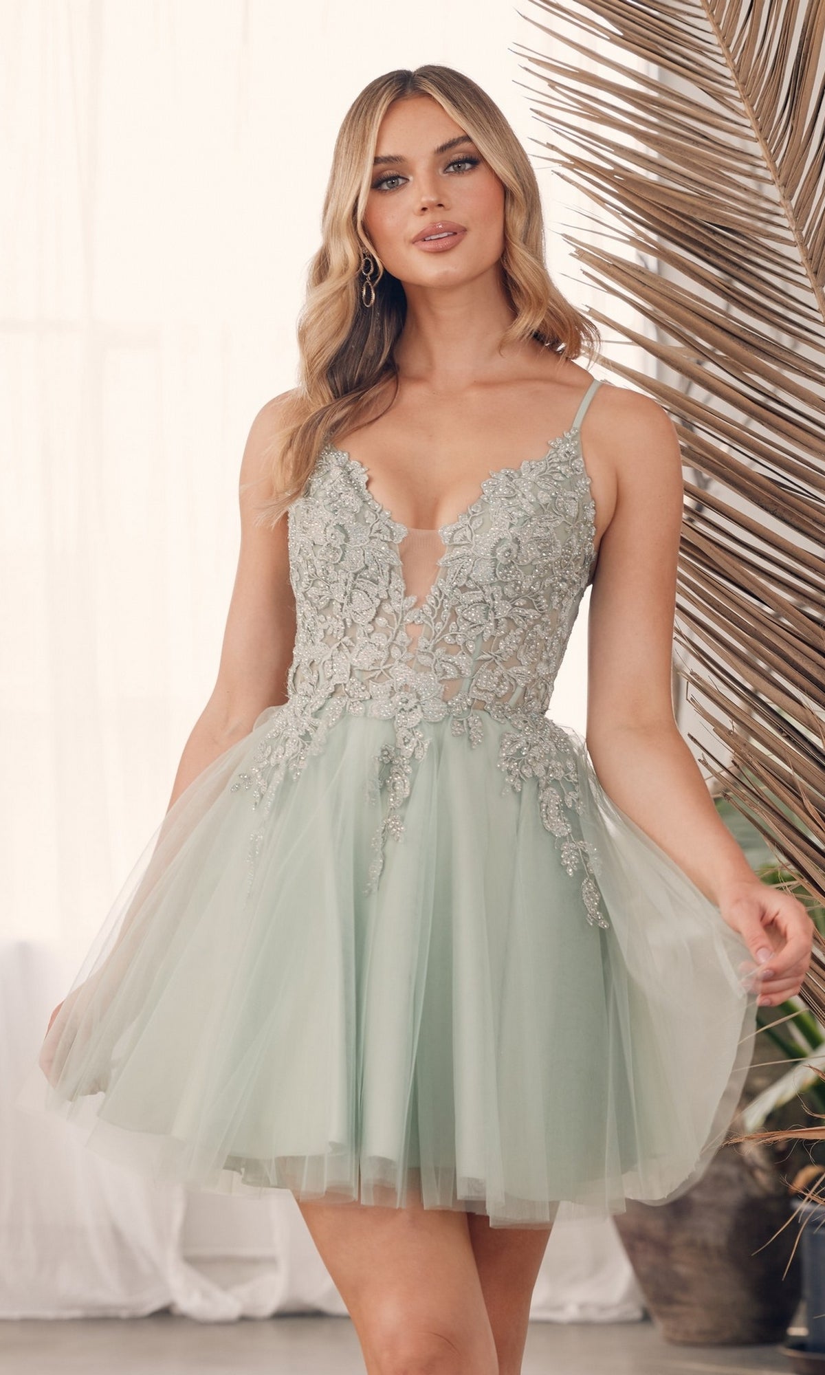 A woman in the Babydoll Lace-Bodice Short Homecoming Dress G785, featuring a pale green tulle skirt and sheer floral-lace bodice, stands indoors by dried palm leaves.