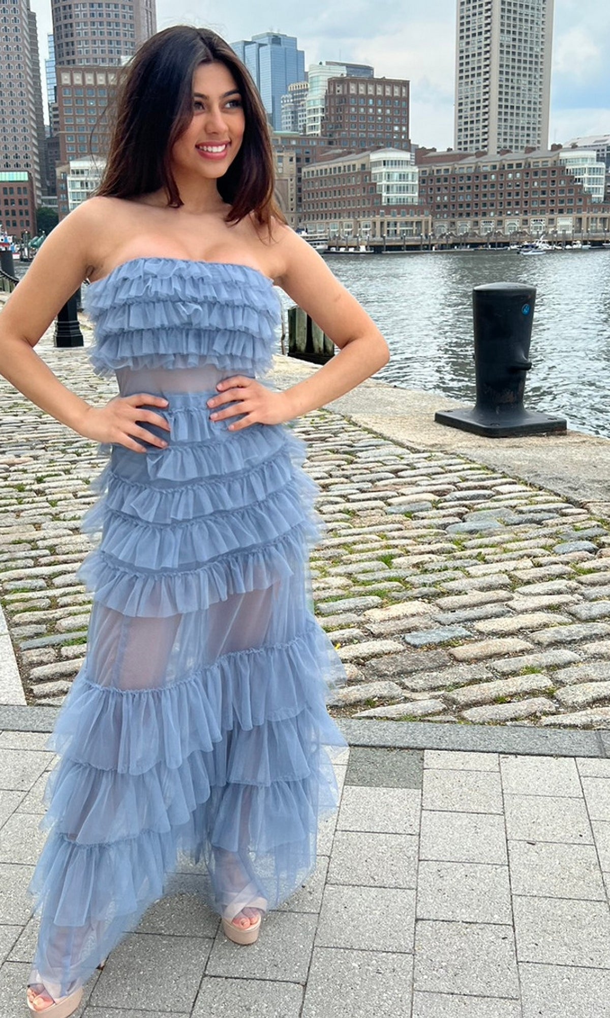 A woman stands on a waterfront promenade in the Sheer Strapless Long Ruffled Prom Dress Georgia with city buildings in the background.