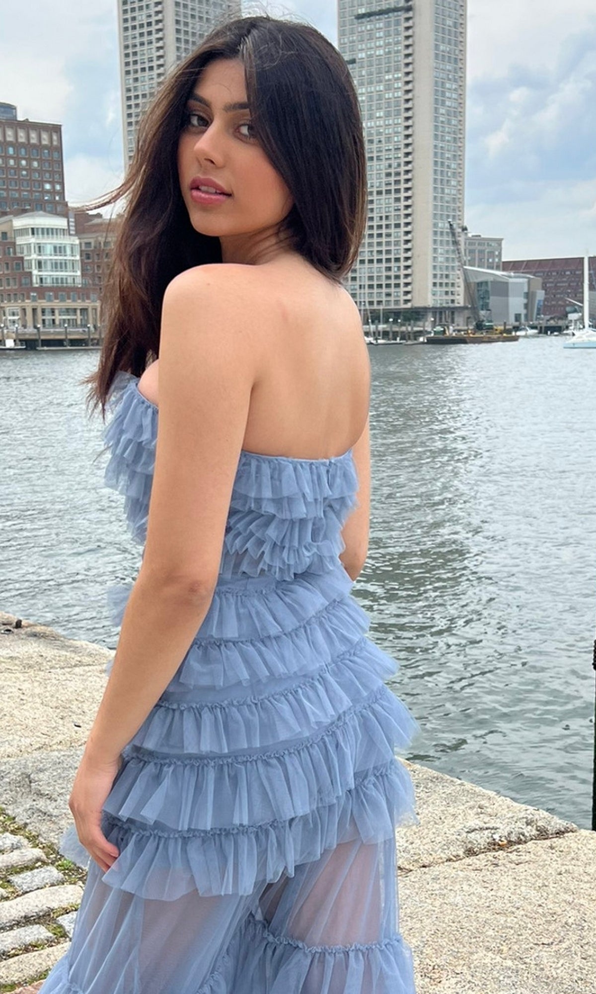 A woman in the Sheer Strapless Long Ruffled Prom Dress Georgia stands by a waterfront, with tall buildings in the background.