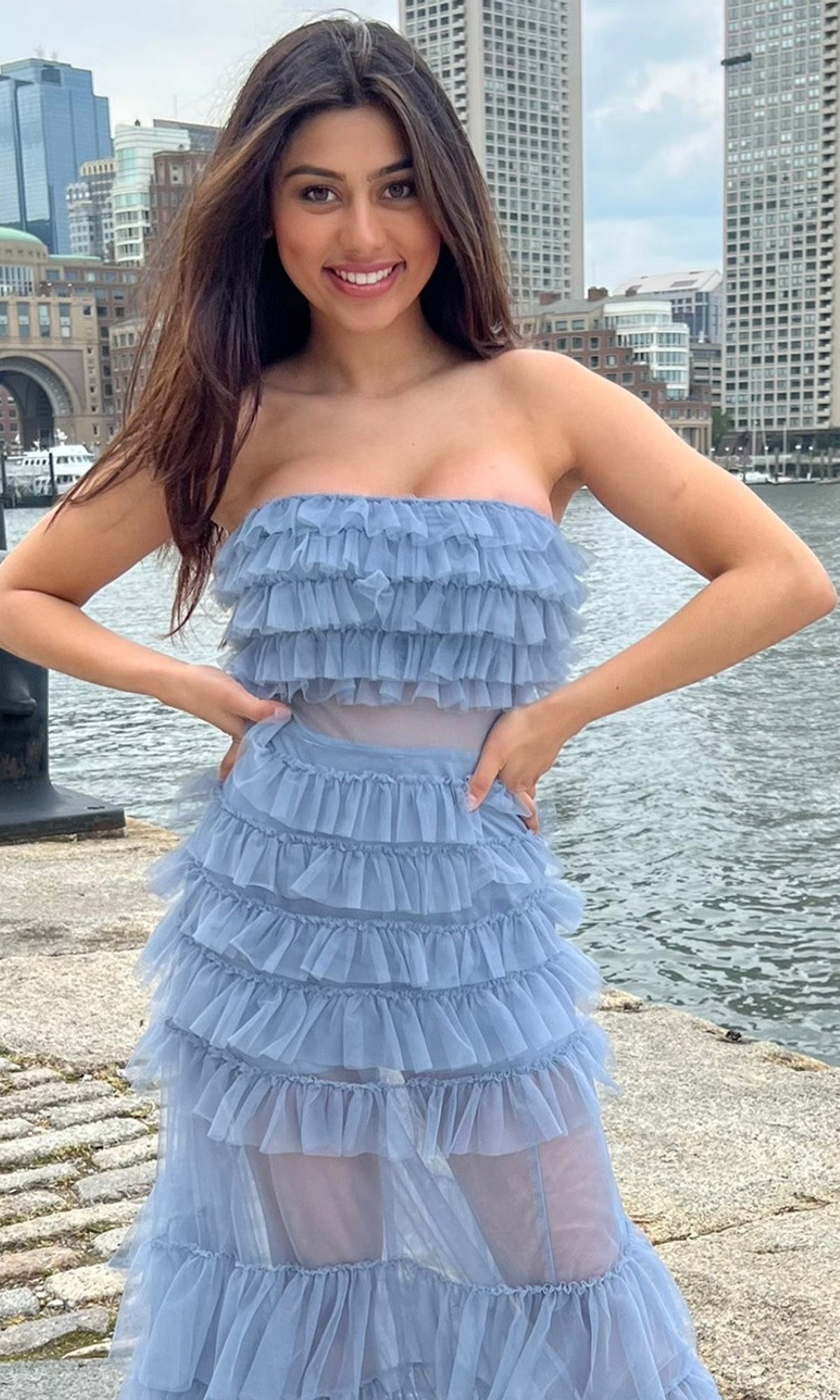 A woman stands by the waterfront in the Sheer Strapless Long Ruffled Prom Dress Georgia, its light blue layers flowing elegantly, with tall city buildings in the background.