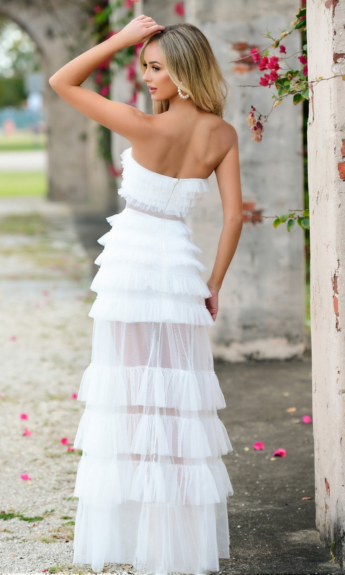 A woman stands outdoors before stone arches, wearing the Sheer Strapless Long Ruffled Prom Dress Georgia and glancing over her shoulder. Pink flowers bloom on a nearby vine.
