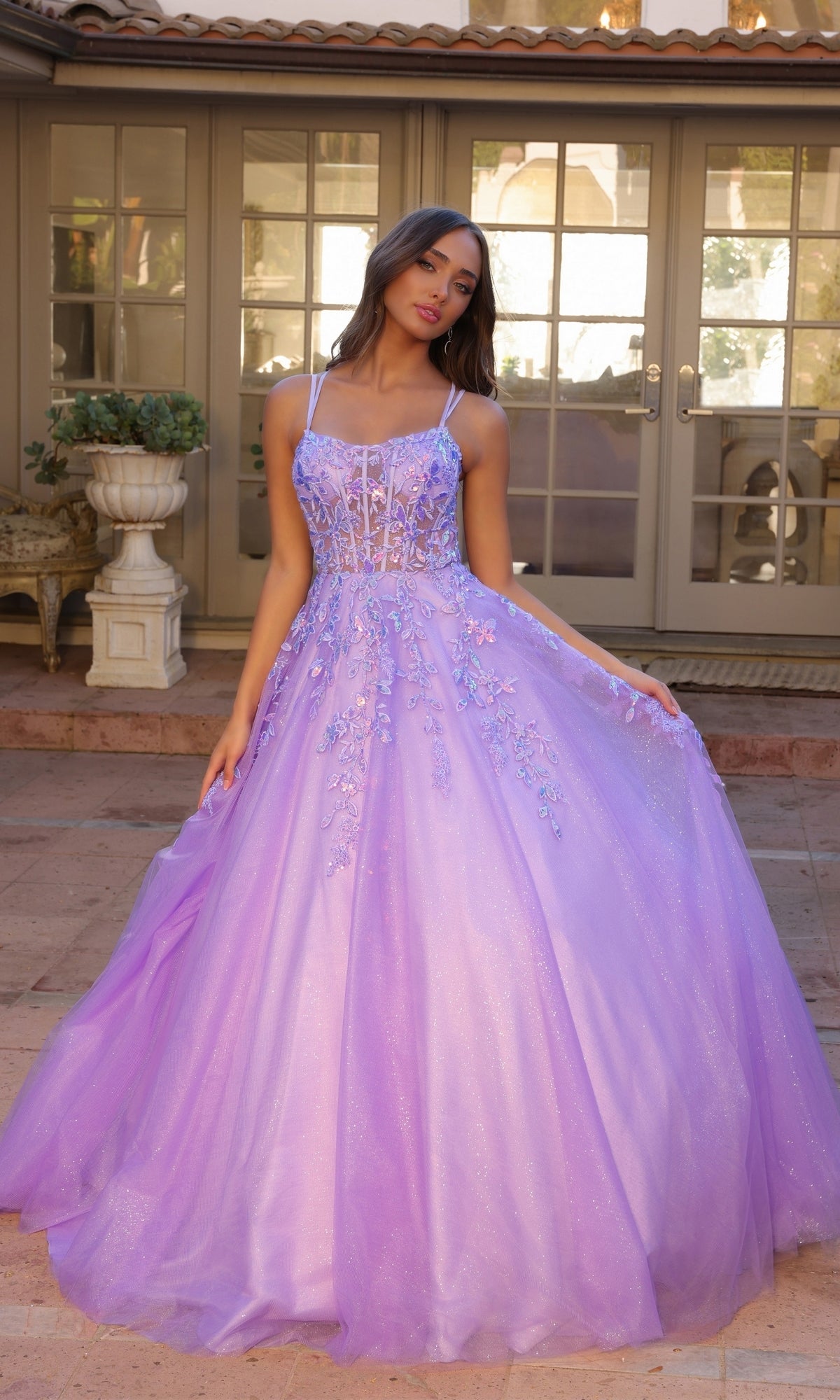 A young woman stands outside, gracefully holding out the skirt of the Glitter-Tulle Long Prom Ball Gown H1271—a lavender ball gown featuring glitter tulle, floral embellishments, and a sheer bodice.