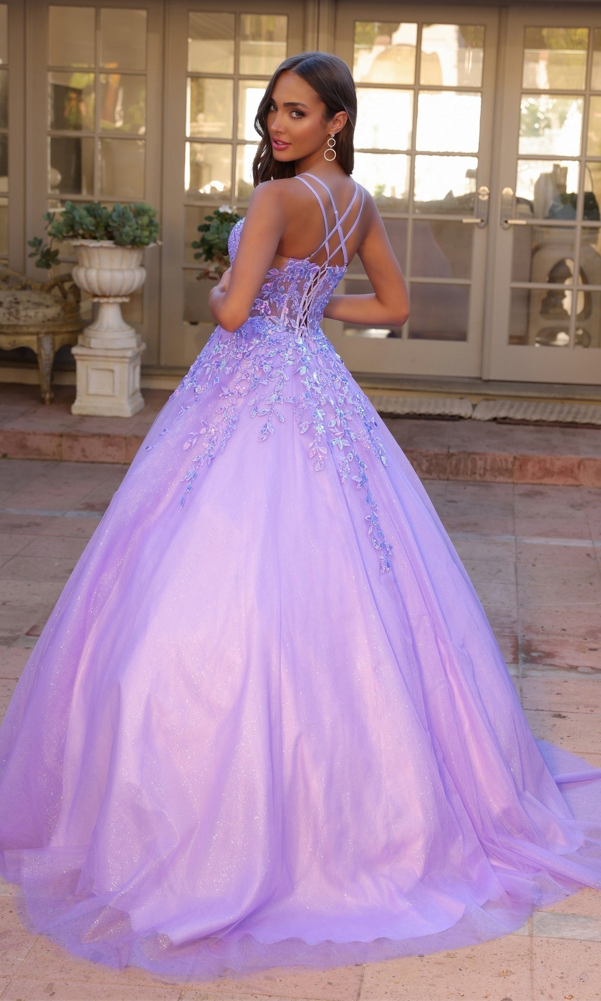 A woman in the Glitter-Tulle Long Prom Ball Gown H1271 with floral accents and a strappy back stands on a patio, looking over her shoulder.