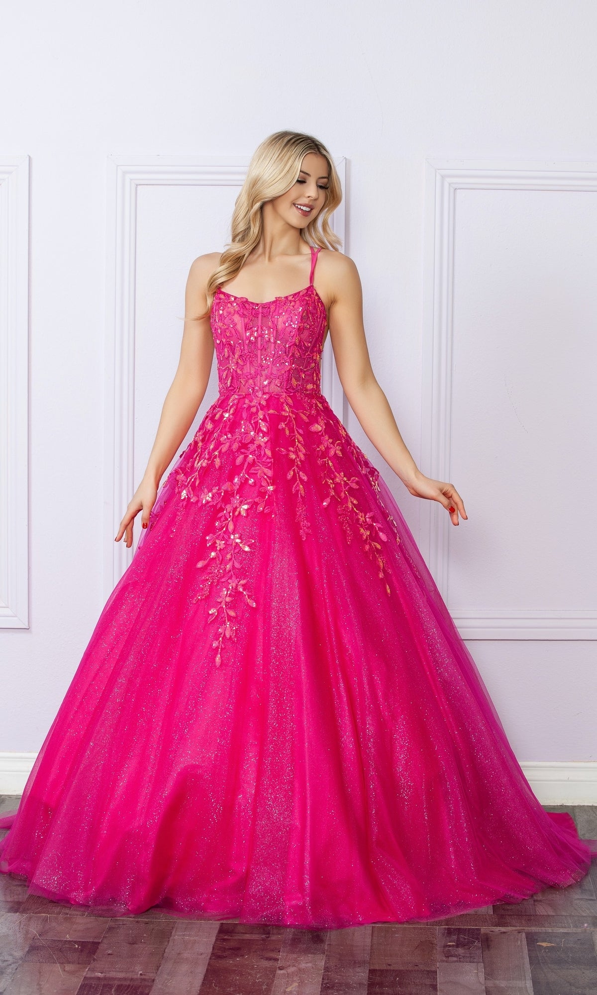 A woman stands against a light wall, smiling to the side in the Glitter-Tulle Long Prom Ball Gown H1271—a bright pink, floor-length dress with floral embellishments.