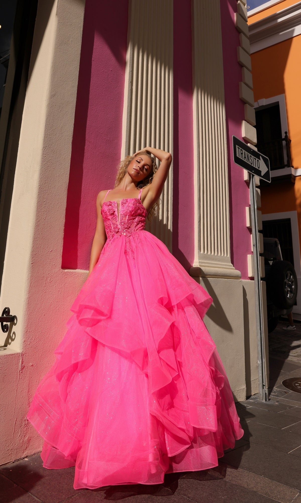 A woman in the Lace-Up Long Tiered Prom Ball Gown H1351, a bright pink strapless dress with tulle ruffles, stands by a pink and cream building near a street sign reading