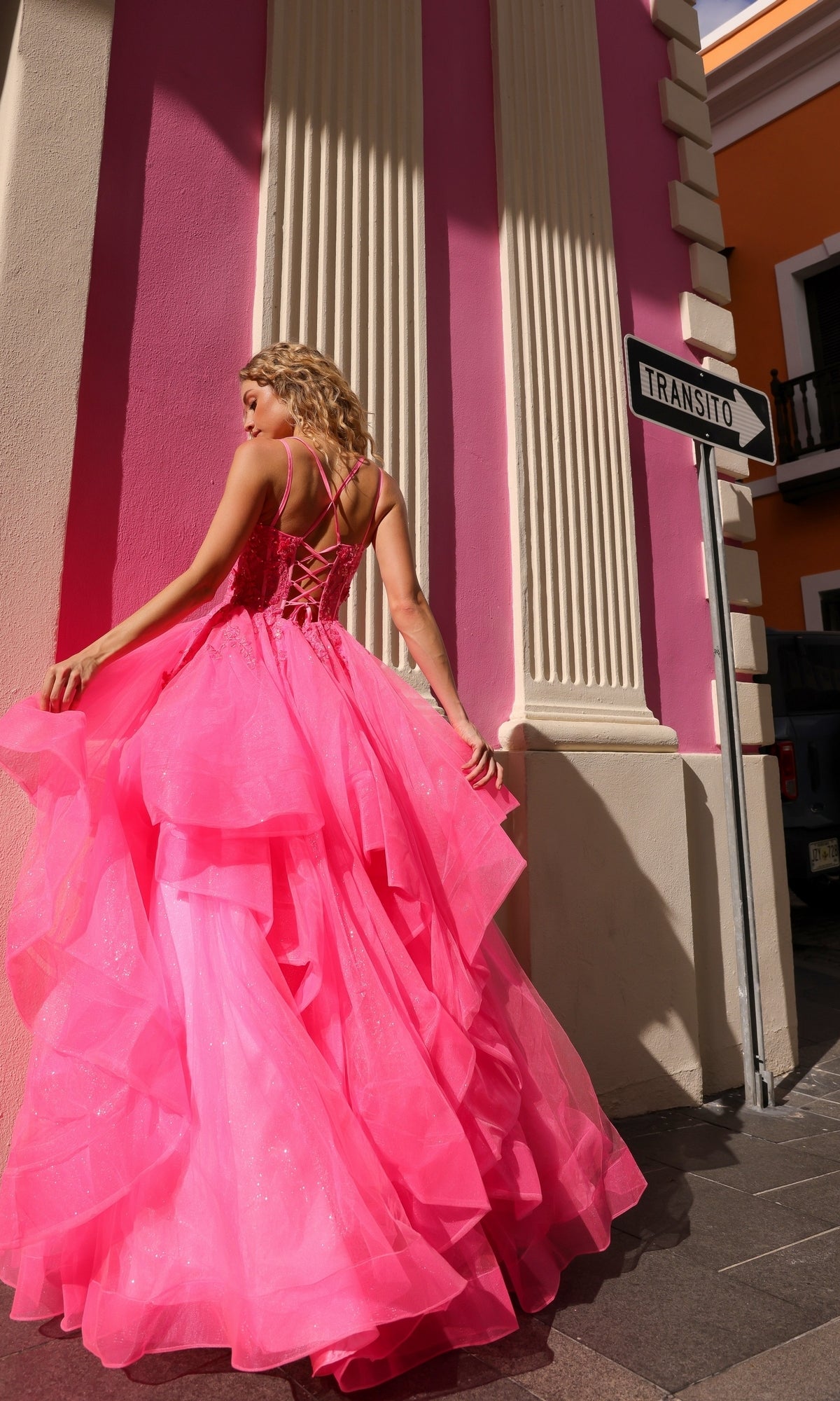 A woman in the Lace-Up Long Tiered Prom Ball Gown H1351, a bright pink dress with tulle ruffles, stands near a