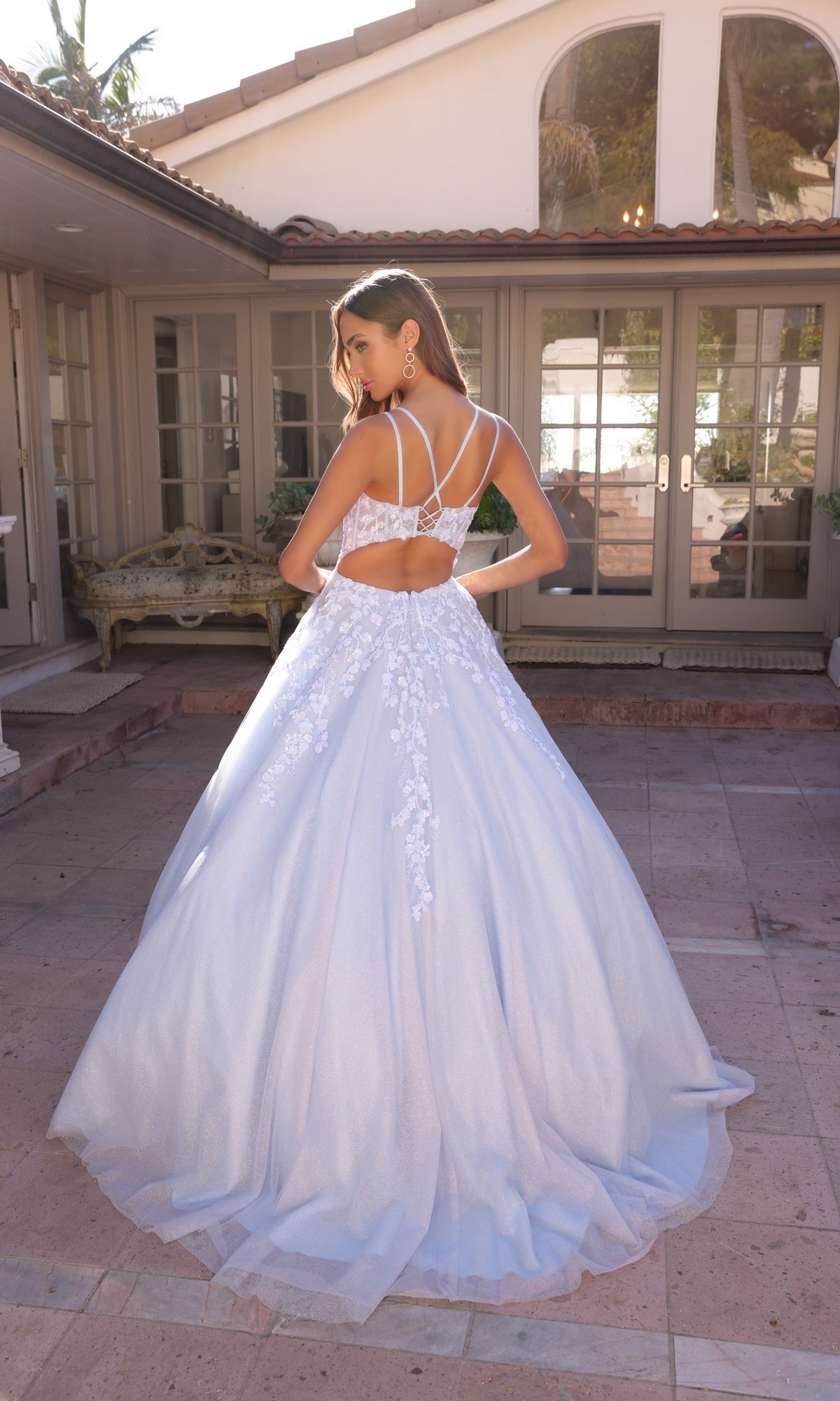 A woman in the Long Corset Glitter Prom Ball Gown H1357, featuring a lace bodice and open strappy back, stands on a stone patio in front of a house with large windows.