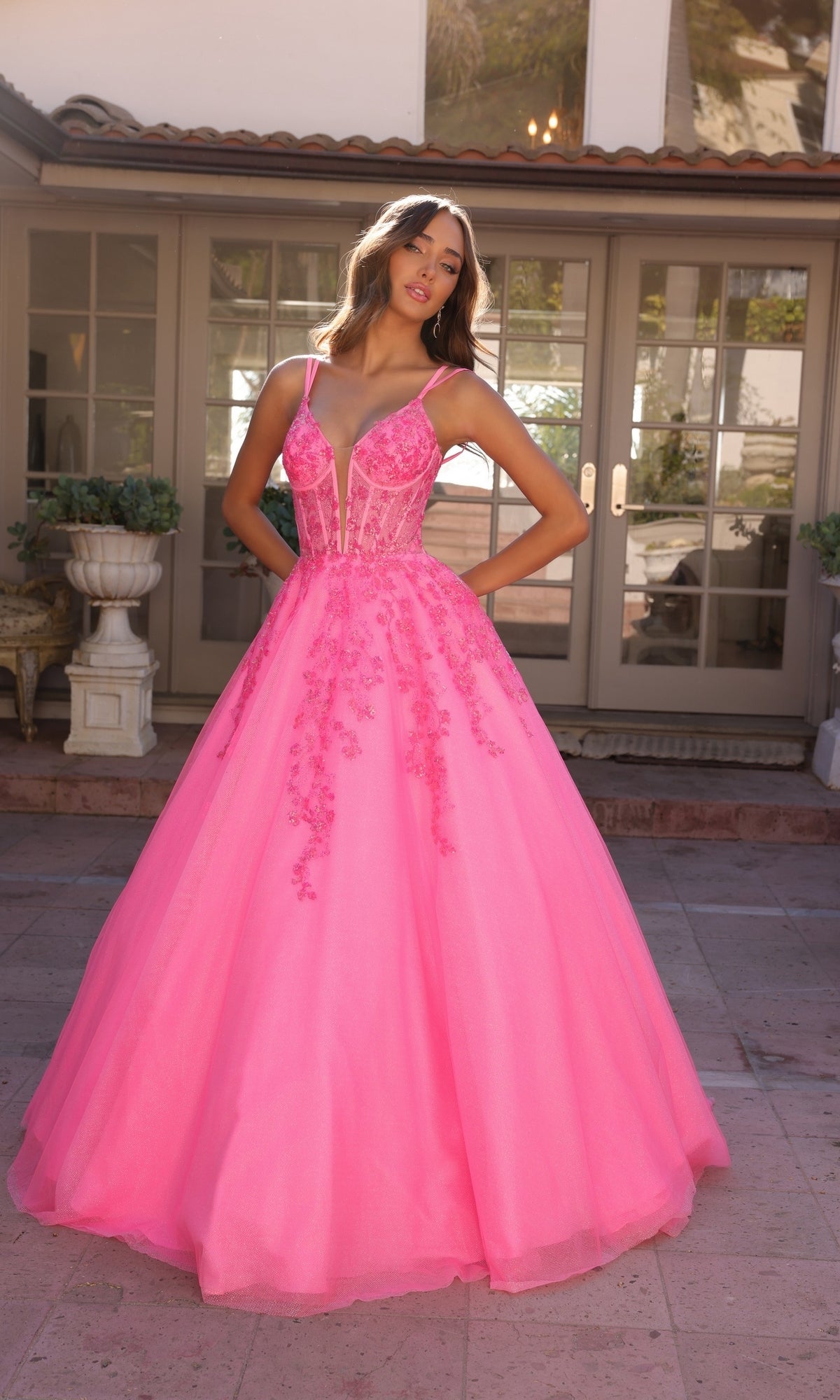 A woman poses gracefully outside glass-paneled doors wearing the Long Corset Glitter Prom Ball Gown H1357, a vibrant pink evening dress with floral appliqué and sheer bodice detailing.