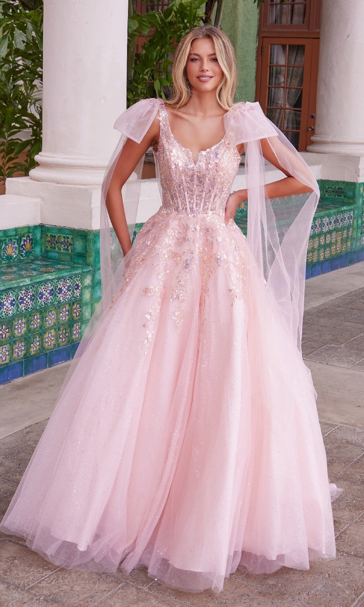 A woman stands outdoors by a tiled wall and white columns, wearing the Nox Anabel H1680 Shoulder-Bows Long Prom Dress—a light pink ball gown with sparkling details, sheer overlays, cap sleeves, and elegant shoulder bows.