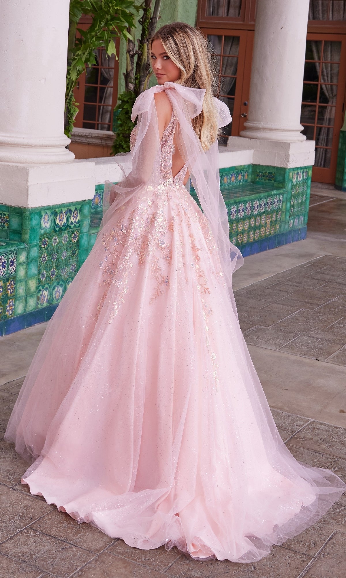 A woman in the Nox Anabel H1680 Shoulder-Bows Long Prom Dress—a light pink, floor-length, backless gown with tulle and sequins—stands outdoors near tiled walls and pillars, gazing over her shoulder.