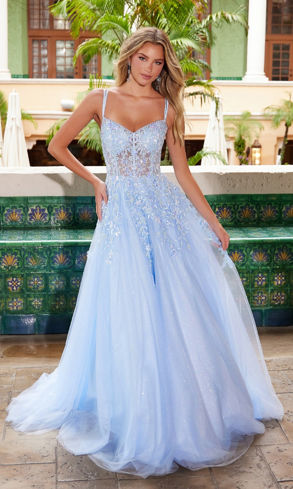 A woman stands outdoors in the Long Glitter-Tulle Prom Dress: Nox Anabel H1682, a light blue, floor-length gown with floral embroidery and a sheer bodice. Ornate green tiles and palm trees are visible in the background.