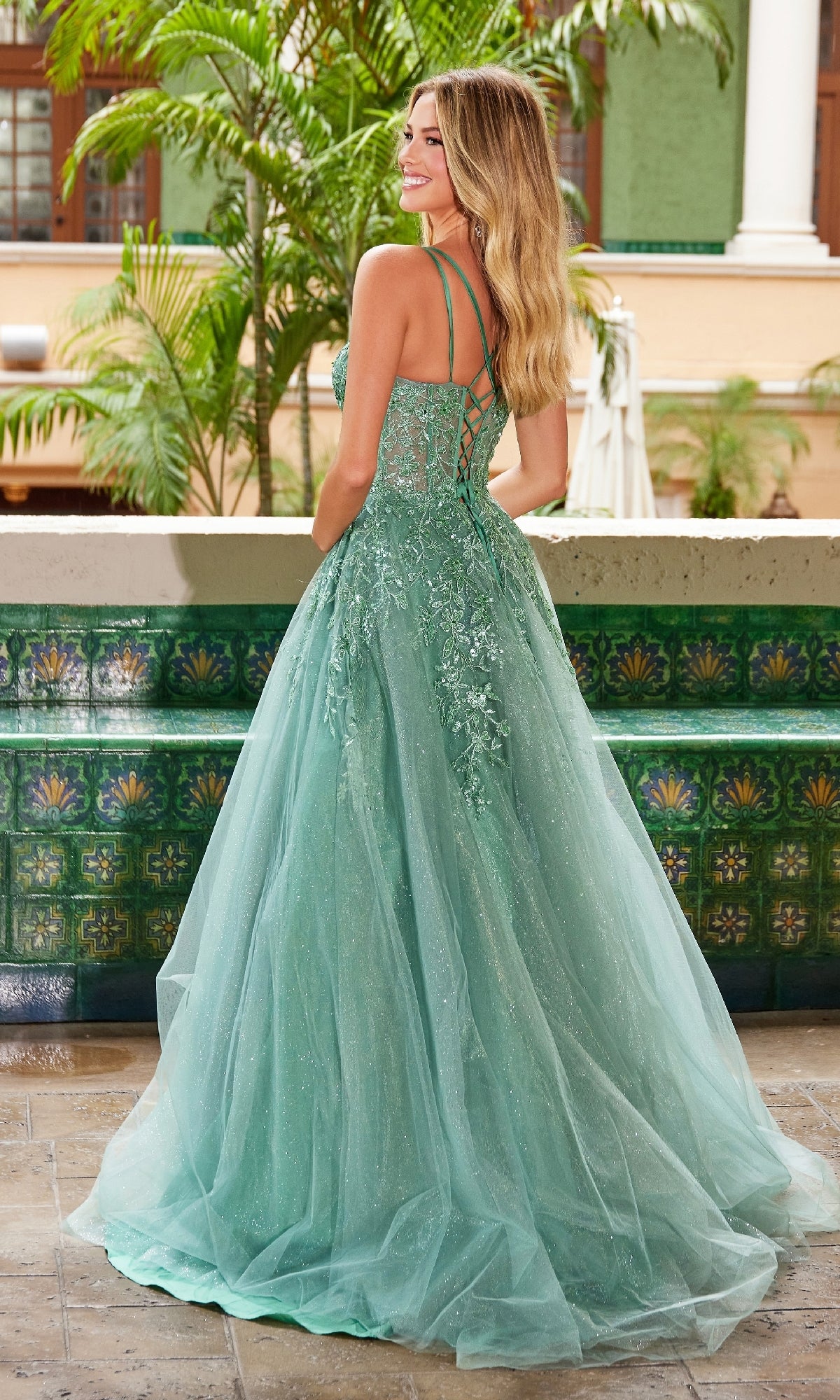 A woman stands outdoors near green tiles and plants, wearing the Nox Anabel H1682 Long Glitter-Tulle Prom Dress in light green, featuring a floor-length design with lace-up detail and floral embroidery.