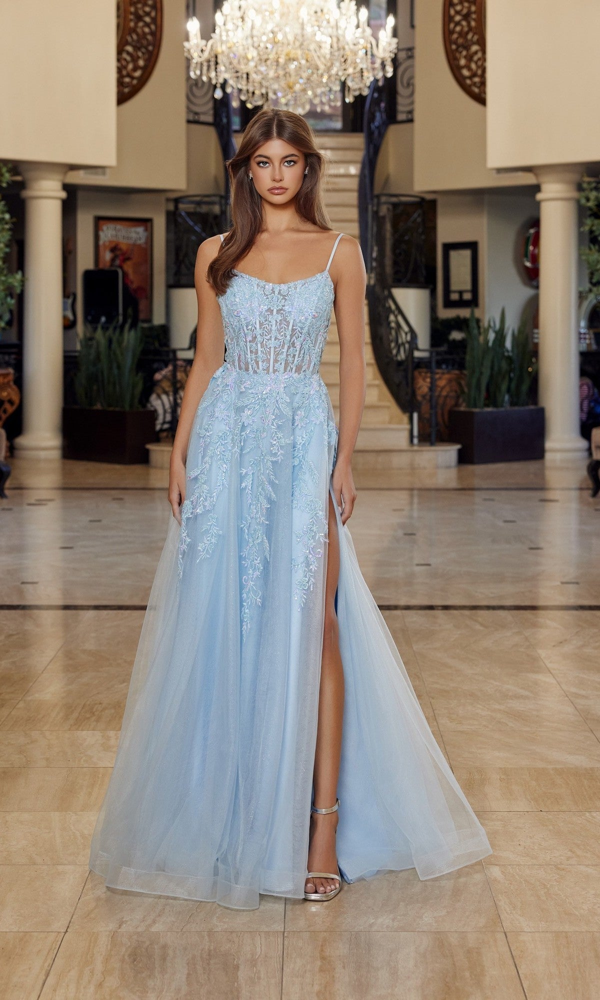 Indoors, a woman wears the blue Nox Anabel H1886 long prom dress featuring a sleeveless design, tulle A-line skirt, sheer bodice with floral embroidery, and high slit. A chandelier and staircase are visible in the background.