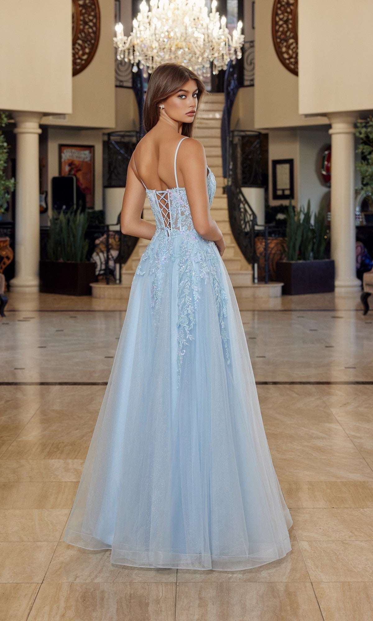 A woman stands in an elegant foyer, facing away from the camera, wearing the Nox Anabel H1886 long prom dress featuring a sleeveless design, tulle A-line skirt, and lace-up back.