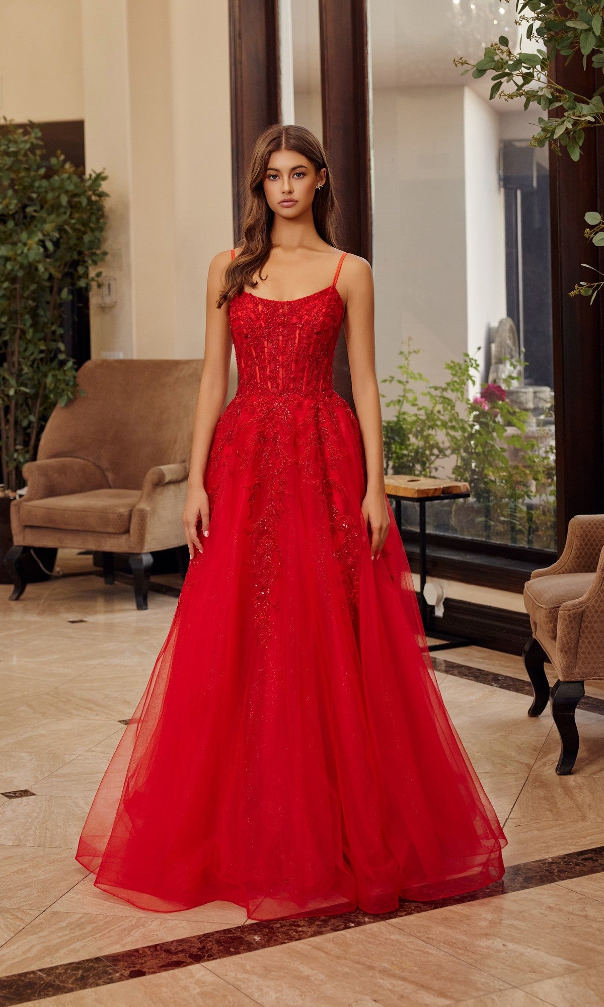 A woman stands indoors wearing the Nox Anabel H1886 long red prom dress, which features thin straps, a fitted bodice, and a flowing tulle A-line skirt. Two armchairs and potted plants are in the background.