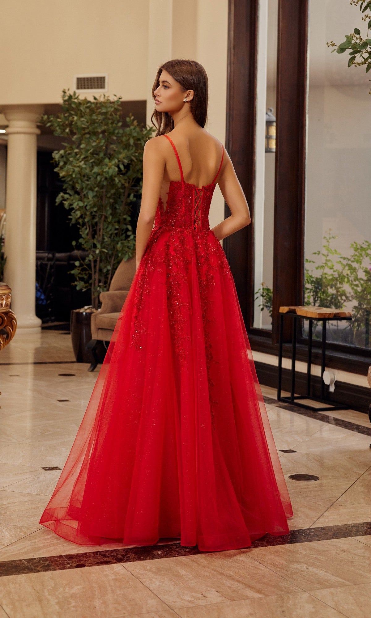 A woman in the Nox Anabel H1886 long prom dress, featuring a sleeveless design and tulle A-line skirt, stands indoors with her back turned in a spacious room with large windows and marble floors.