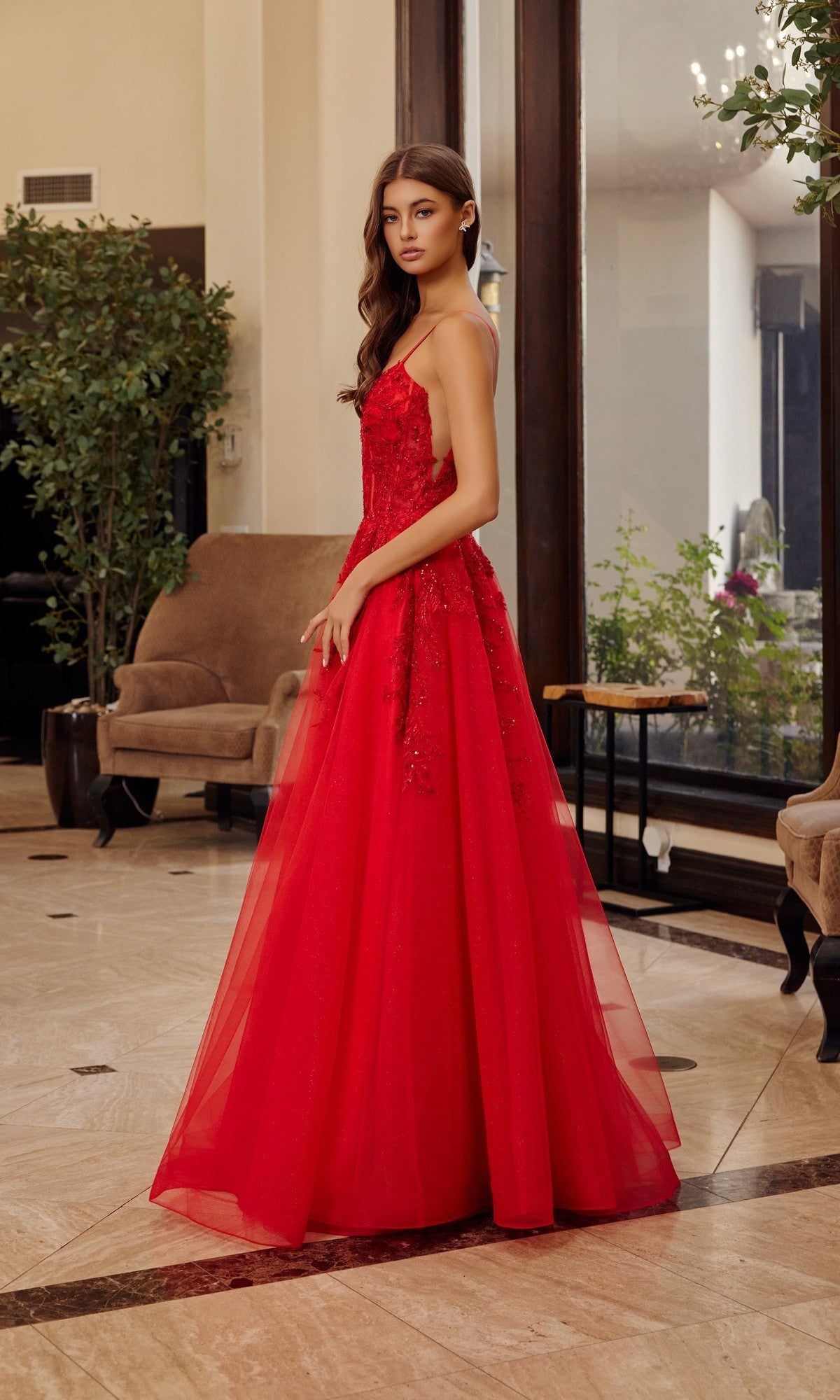 A woman stands indoors wearing the Nox Anabel H1886, a sleeveless long prom dress with a tulle A-line skirt and floral embroidery, posing beside a brown armchair and large windows with greenery outside.