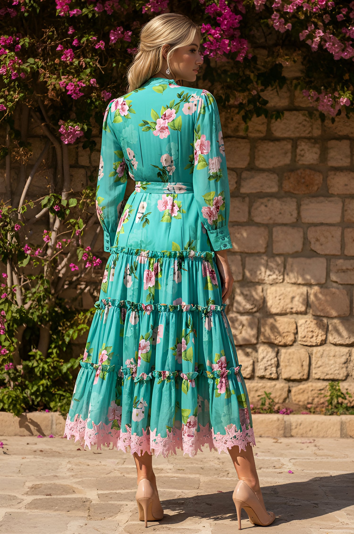 A woman stands outdoors with her back to the camera, wearing the Hazel Tropical Tea Length Casual Dress featuring long sleeves, tiered ruffles, pink lace trim, and a belted waist. She pairs it with nude heels against a stone wall and flowering plants.