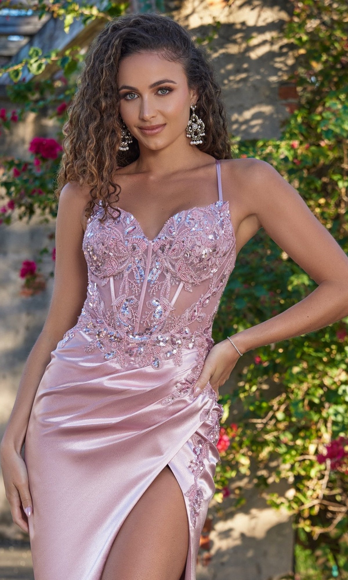 A woman poses outdoors near greenery and flowers wearing the Velvi Lace-Up Sheer-Corset Long Prom Dress Ivy, which features a sweetheart neckline, sequin details, and a thigh-high slit.