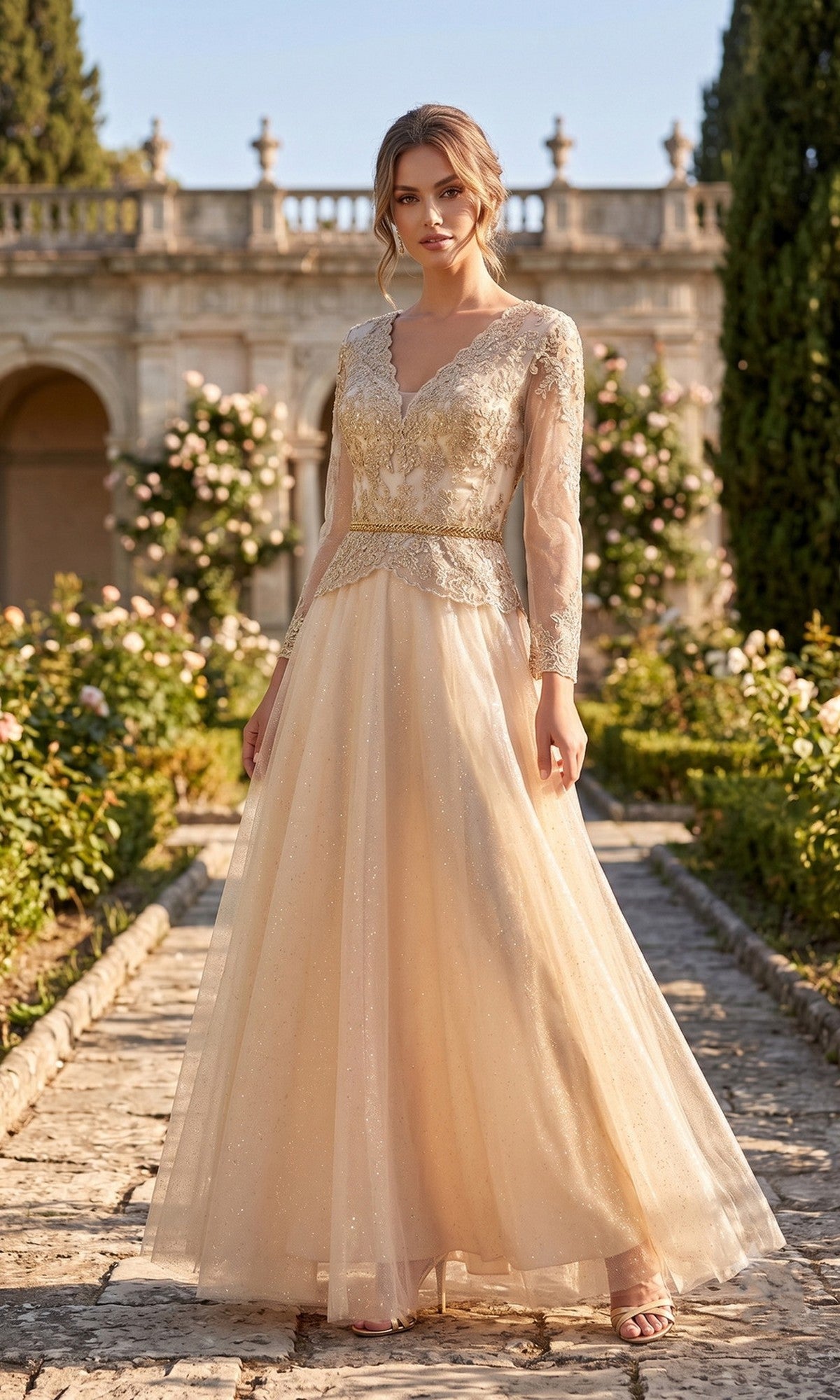 A woman stands outdoors in a garden wearing the Chicas J082 Long Prom Dress in champagne, featuring lace and bead details that accentuate the embellished bodice, creating an elegant look perfect for a stunning mother of the bride. Greenery and a stone building provide a picturesque backdrop.