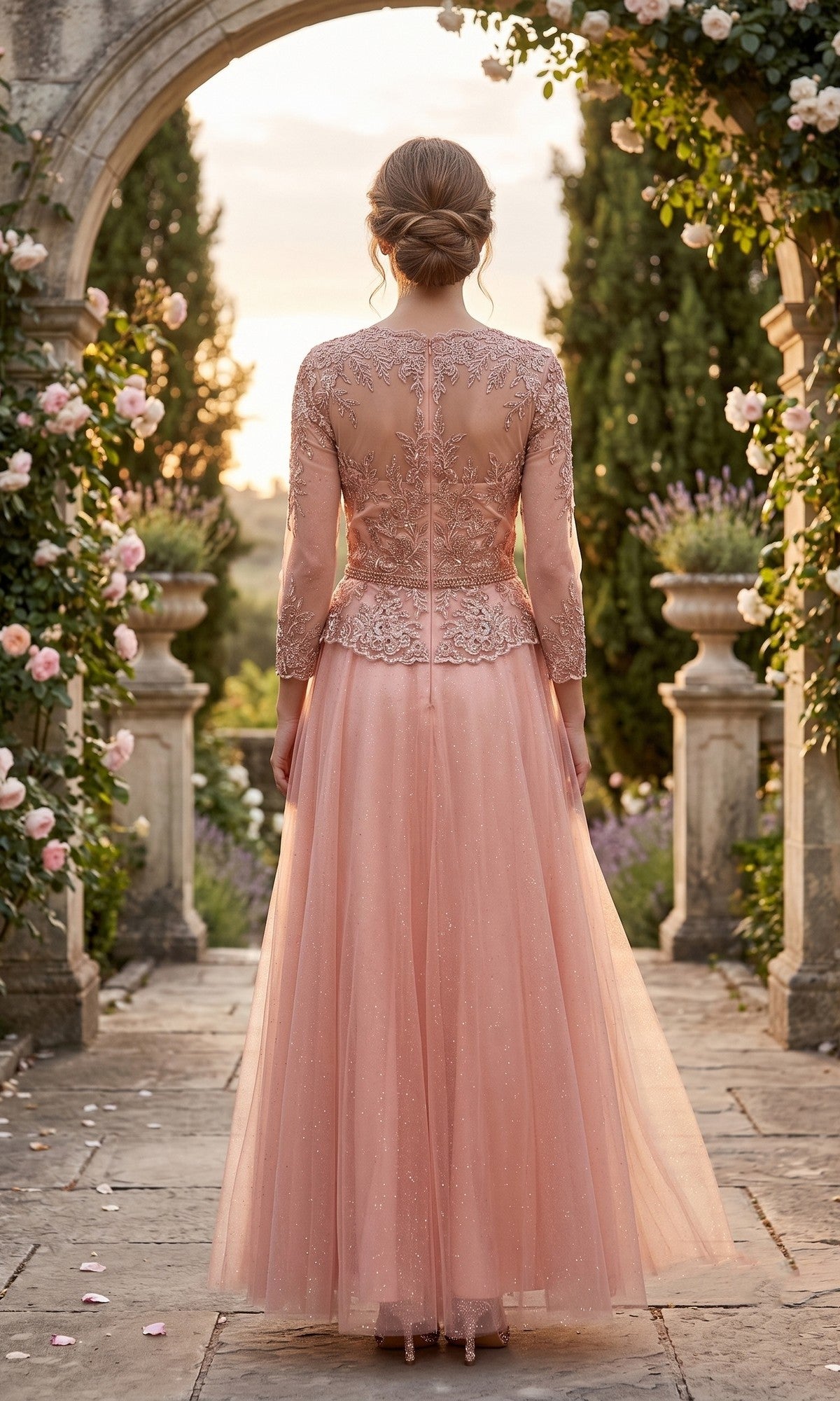 A woman stands outdoors on a stone path, surrounded by flowers and greenery, wearing the Long Prom Dress: Chicas J082—a pink, floor-length gown with intricate embroidery and an embellished bodice.