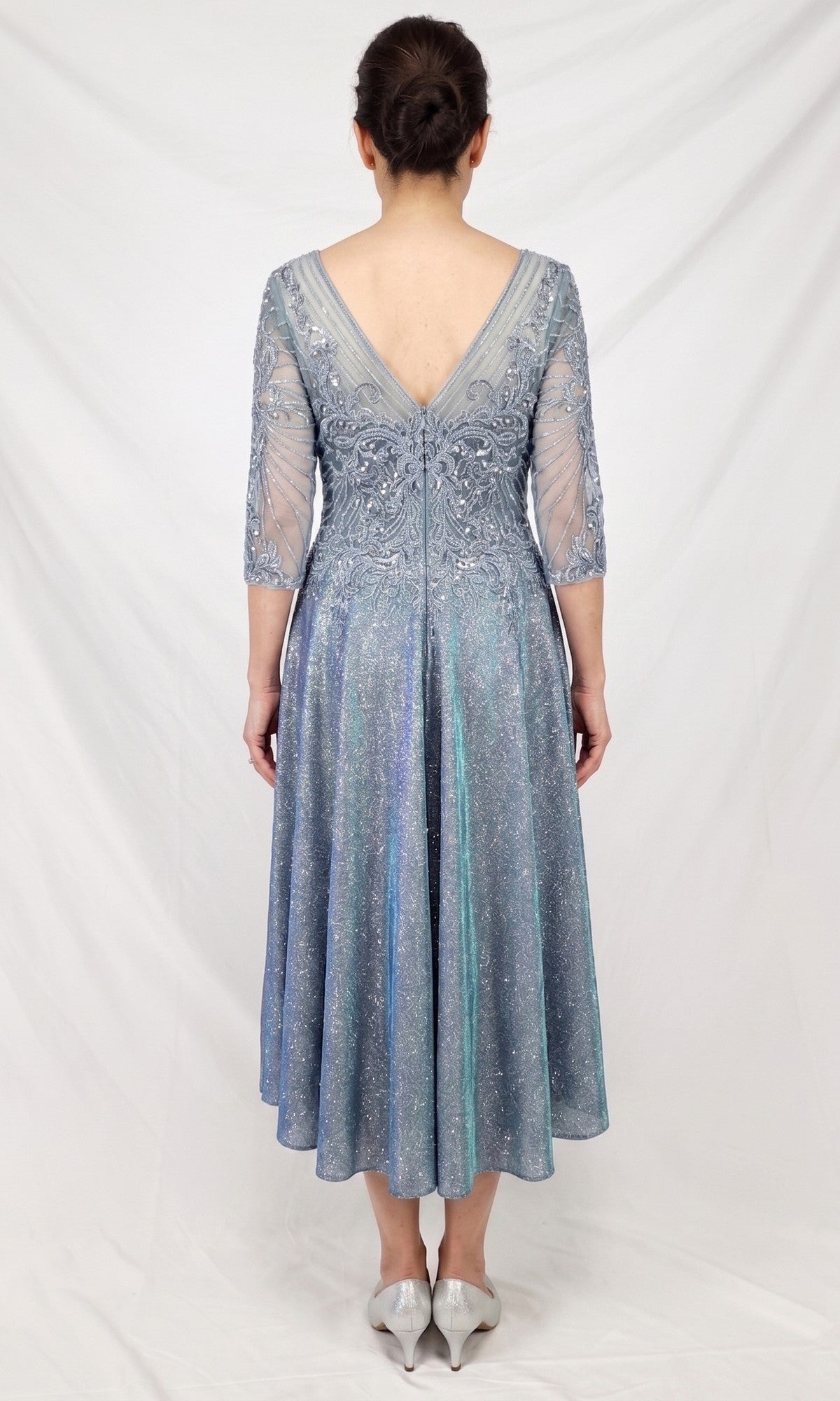 A woman stands facing away, wearing the Modest Midi Wedding Guest Dress: Chicas J152—a silver-blue, knee-length A-line midi dress with a V-back and sheer sleeves—paired with silver shoes against a plain white background, creating a chic and elegant look for any wedding guest.