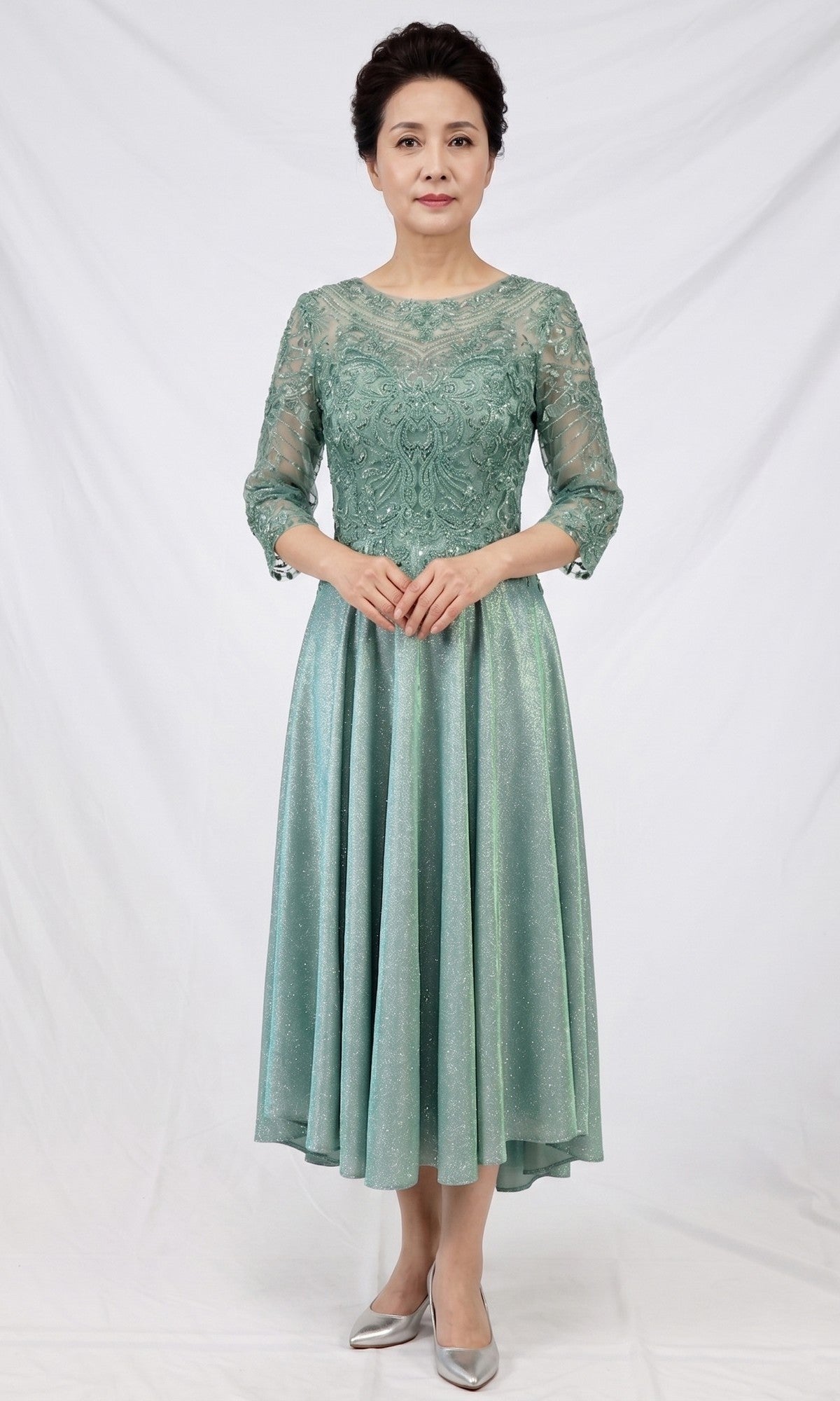 A woman stands in front of a white backdrop wearing the Modest Midi Wedding Guest Dress: Chicas J152 in sage, featuring lace and satin fabric, an A-line midi silhouette with three-quarter sleeves, paired with silver shoes.